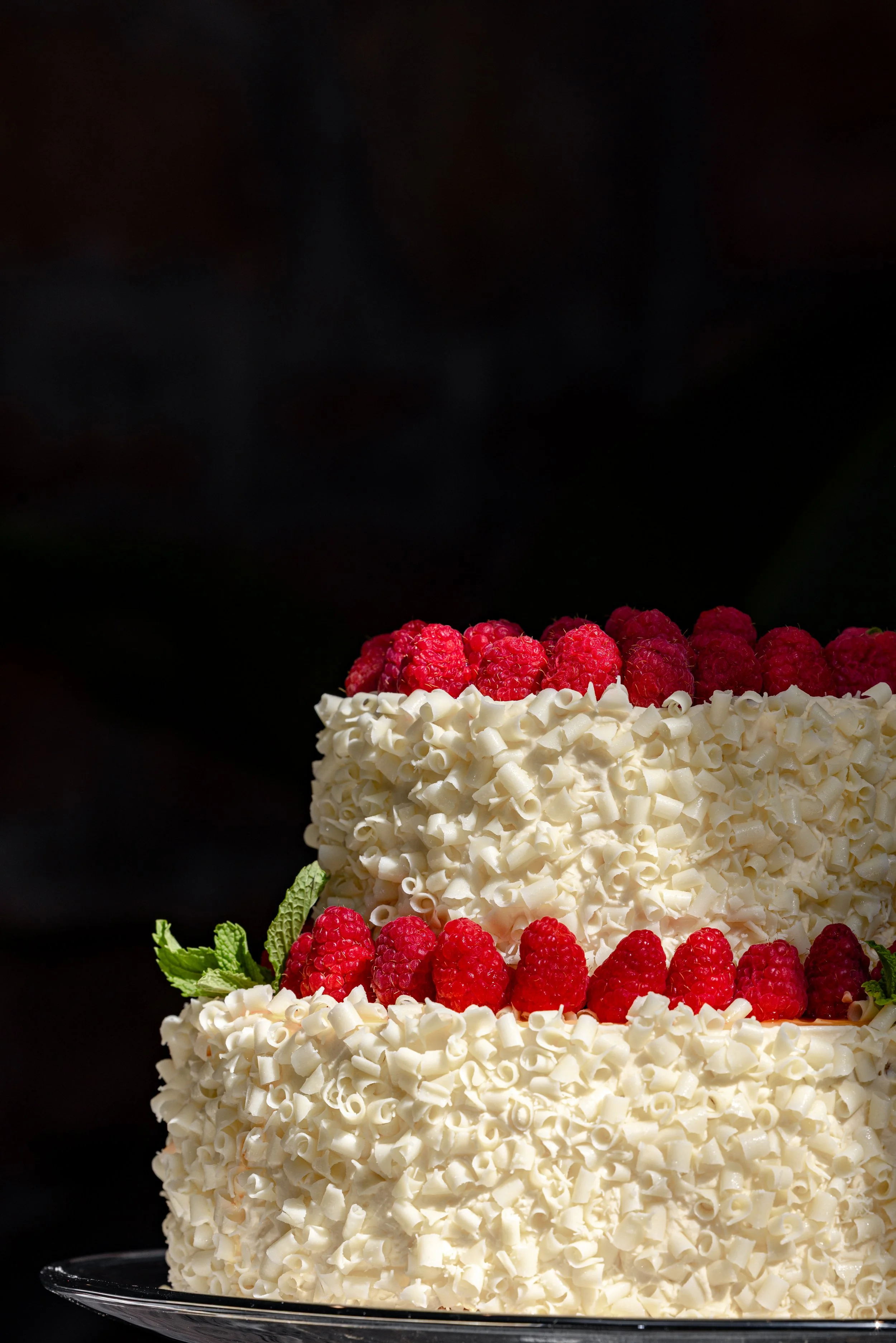A multi-layered cake decorated with white chocolate curls, fresh raspberries, and green mint leaves against a black background.
