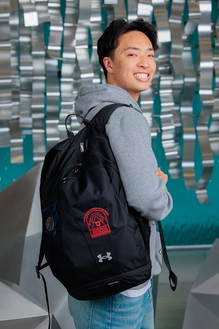 Young man smiling with arms crossed, wearing a gray hoodie, carrying a black backpack, standing indoors in front of a modern metallic sculpture.
