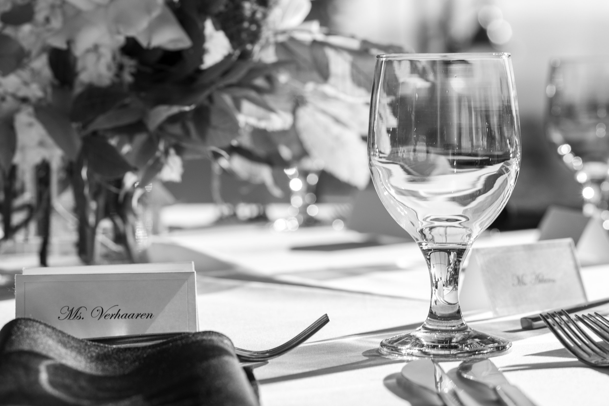 A black and white photo of a table setting with a wine glass, a name card that reads 'Ms. Verhaaren,' some cutlery, and a nearby flower arrangement or centerpiece.