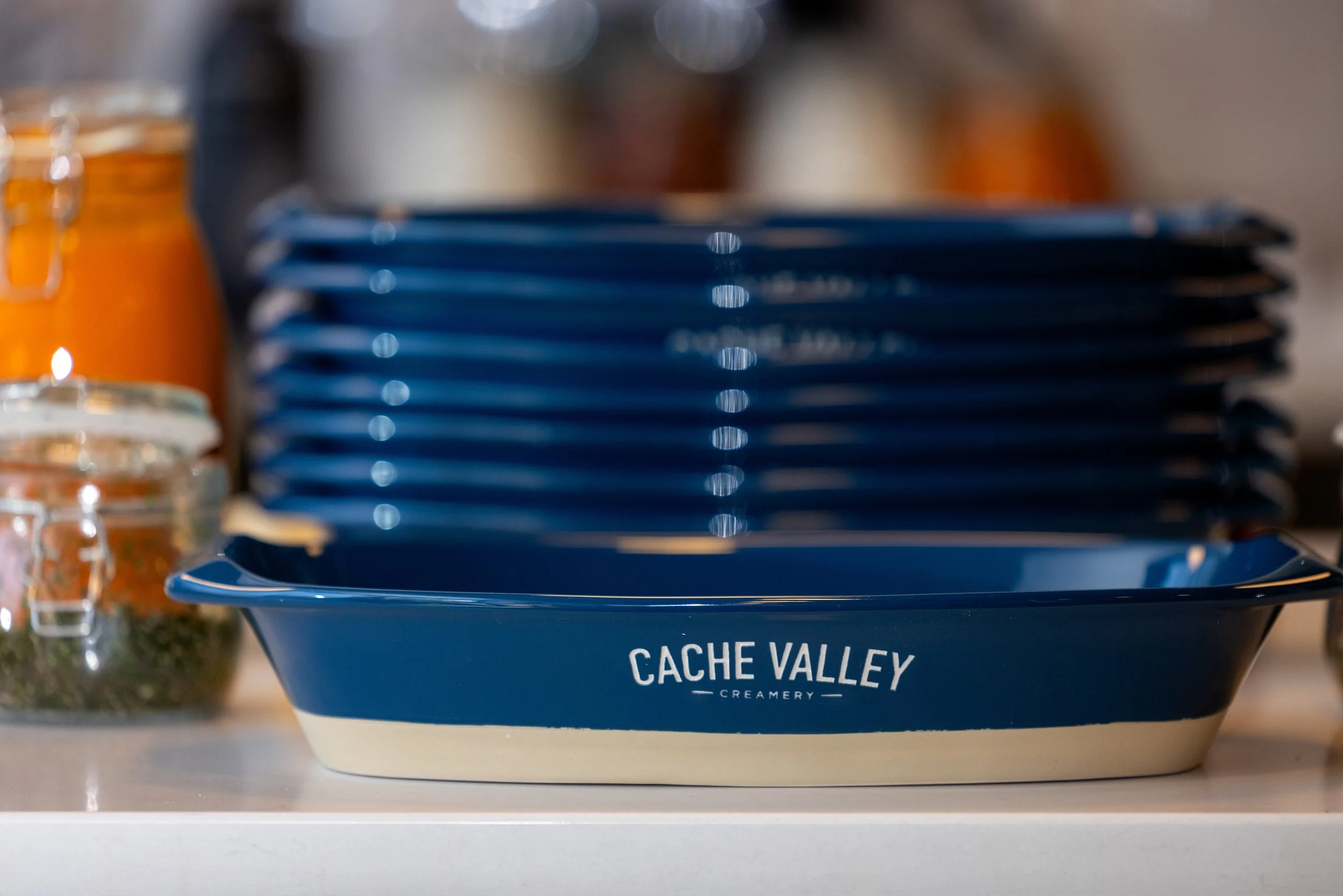 A deep blue bowl with white bottom and inside, labeled "CACHE VALLEY CREAMERY," filled with ice cream. Partially visible ice cream jars are on the left. The background is blurred.