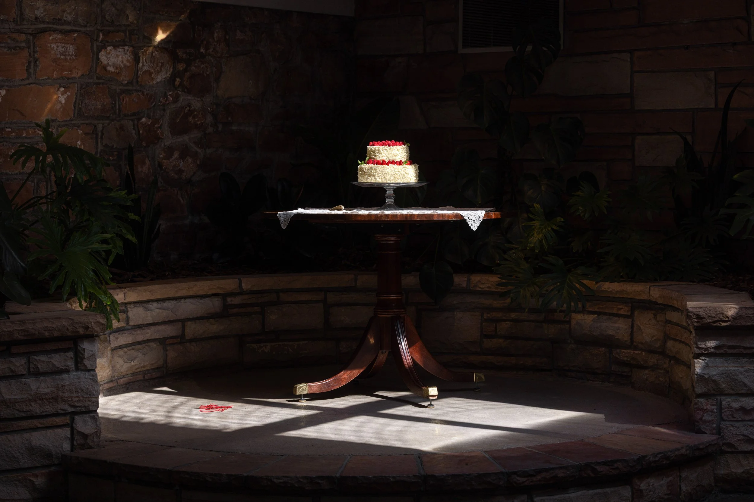 A two-tier white cake decorated with red berries on top, displayed on a glass cake stand on a wooden table with a lace cloth, in a dimly lit room with stone and brick walls and some plants.