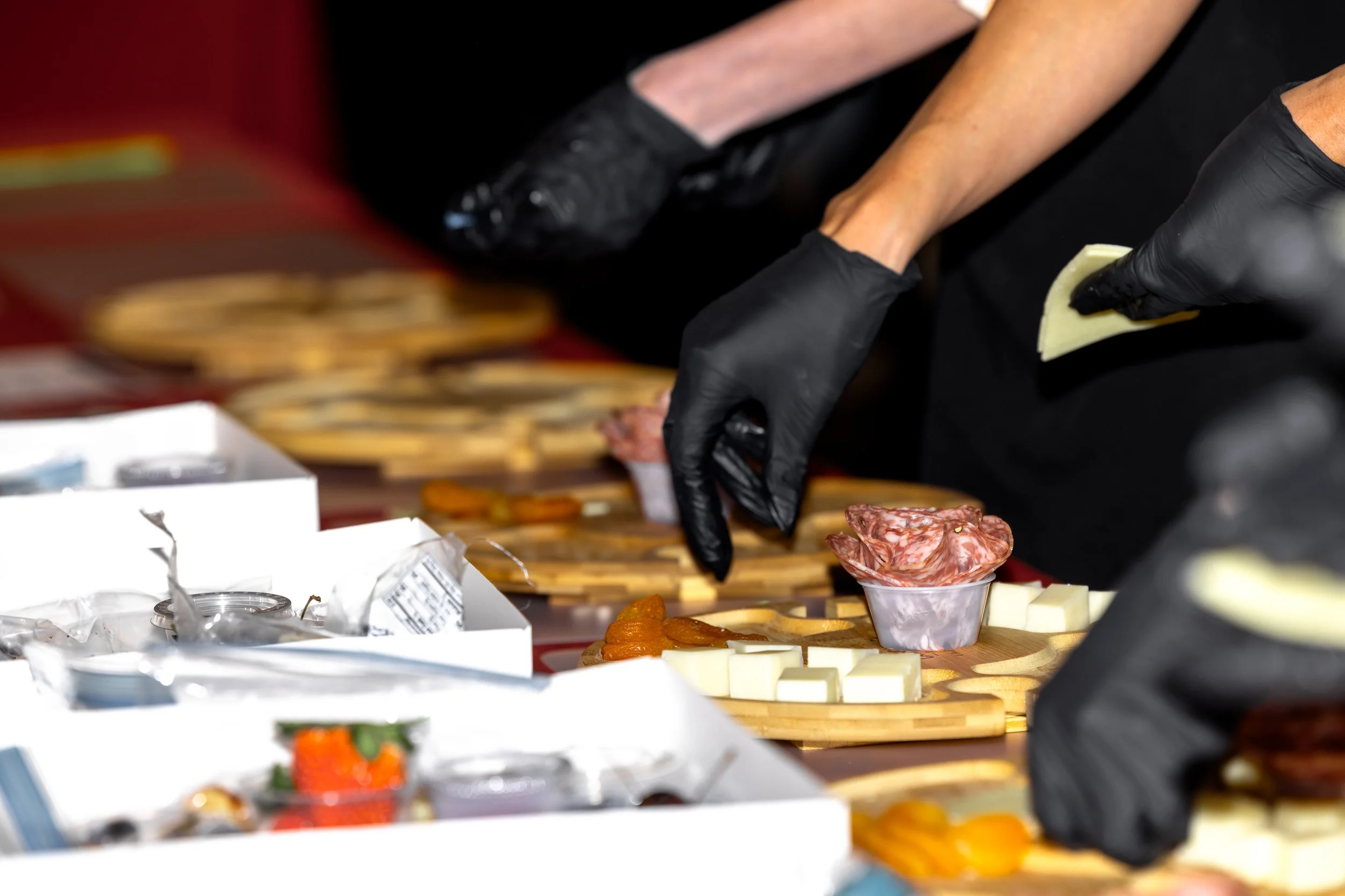Close-up of a person wearing black gloves preparing food, with various cheese and cured meats arranged on a wooden board.