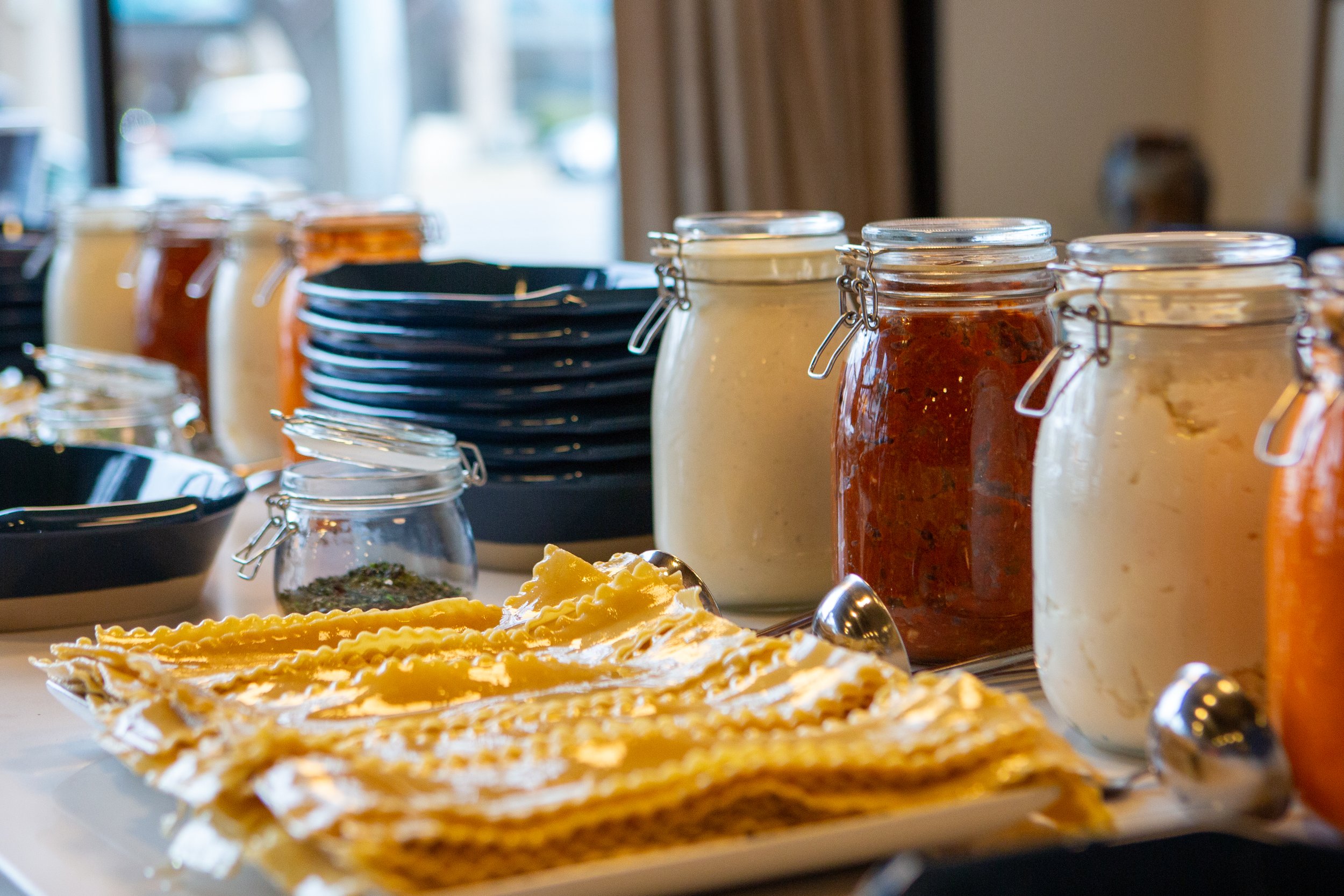 A display of jars of sauces and condiments, black bowls, and a plate of lasagna on a table, with a window and curtains in the background.