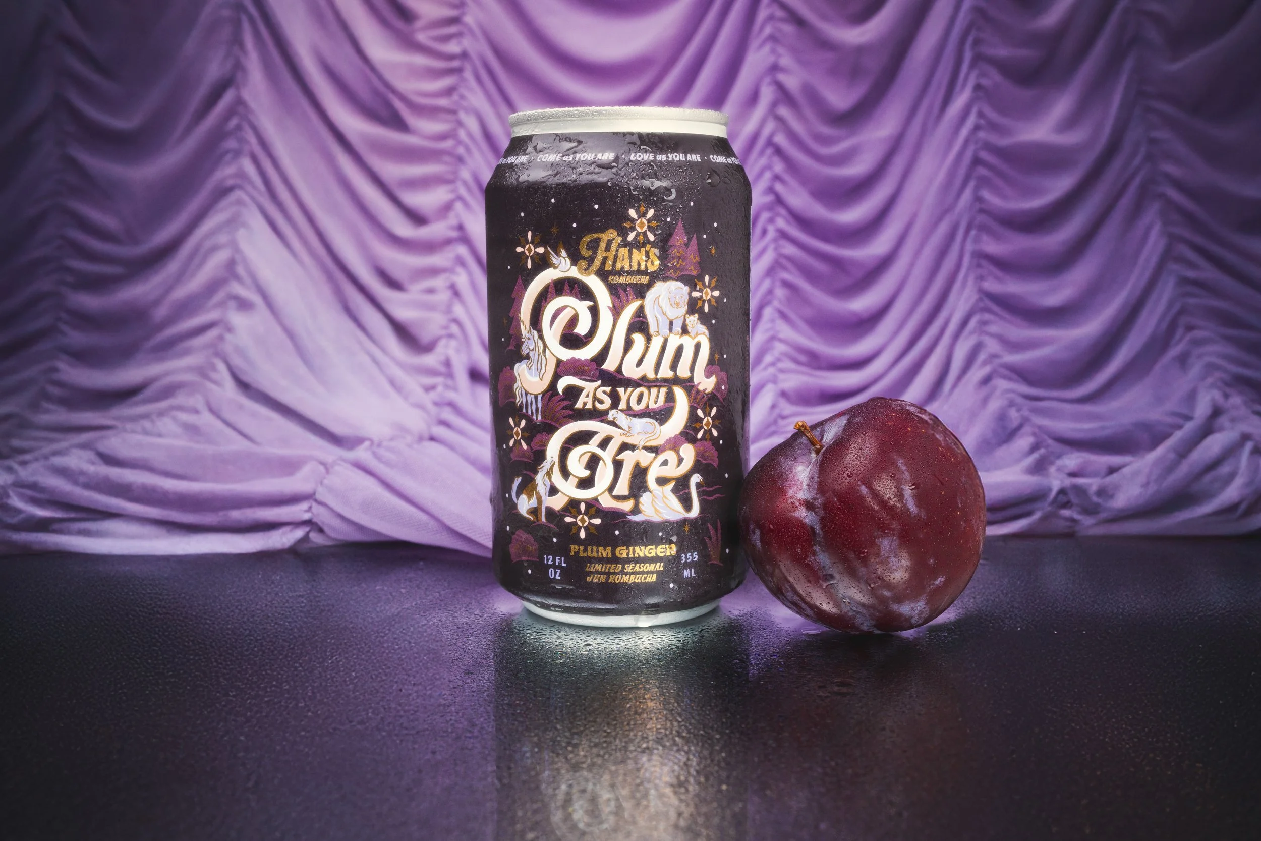A black can of Plum Ginger sparkling beverage with purple and gold graphics and white text, placed on a reflective surface. Next to the can is a whole plum fruit with a dark purple color and a small stem. The background features purple, ruched fabric