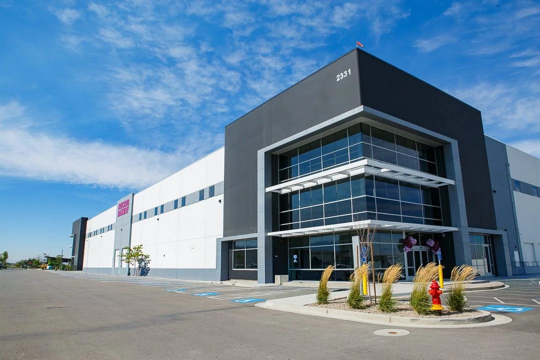 Exterior of a modern large Amazon distribution center with a parking lot in front, clear blue sky, and minimal landscaping.