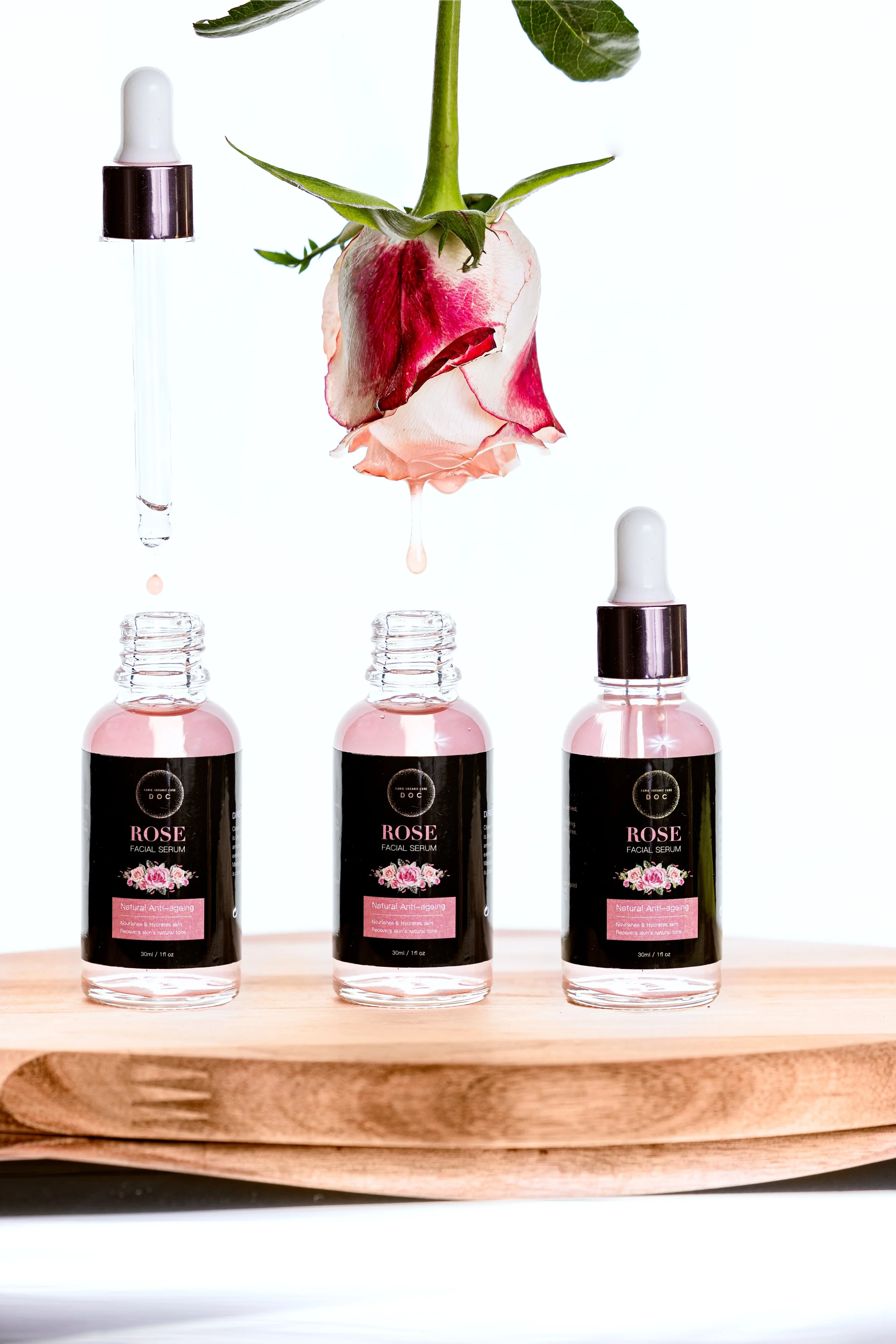 Three bottles of rose facial serum on a wooden surface with a pink and white rose suspended in front of them, and a dropped droplet of serum below the flower.