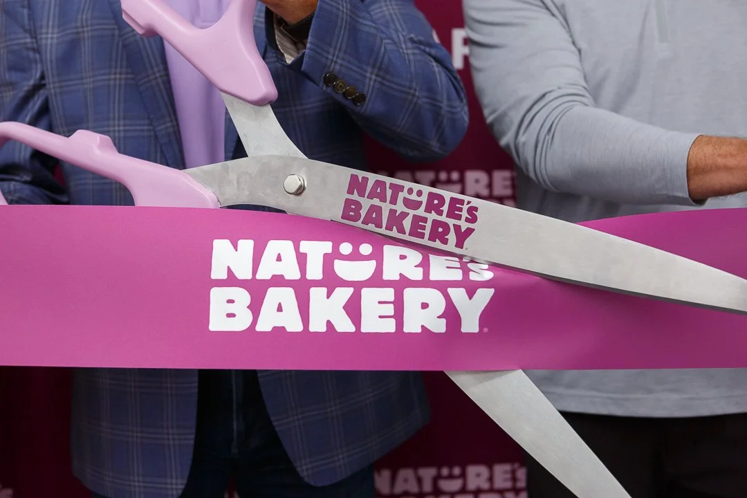 A pair of large scissors cutting a pink ribbon with the words 'Nature's Bakery' in white and purple letters, part of a presentation or opening event.