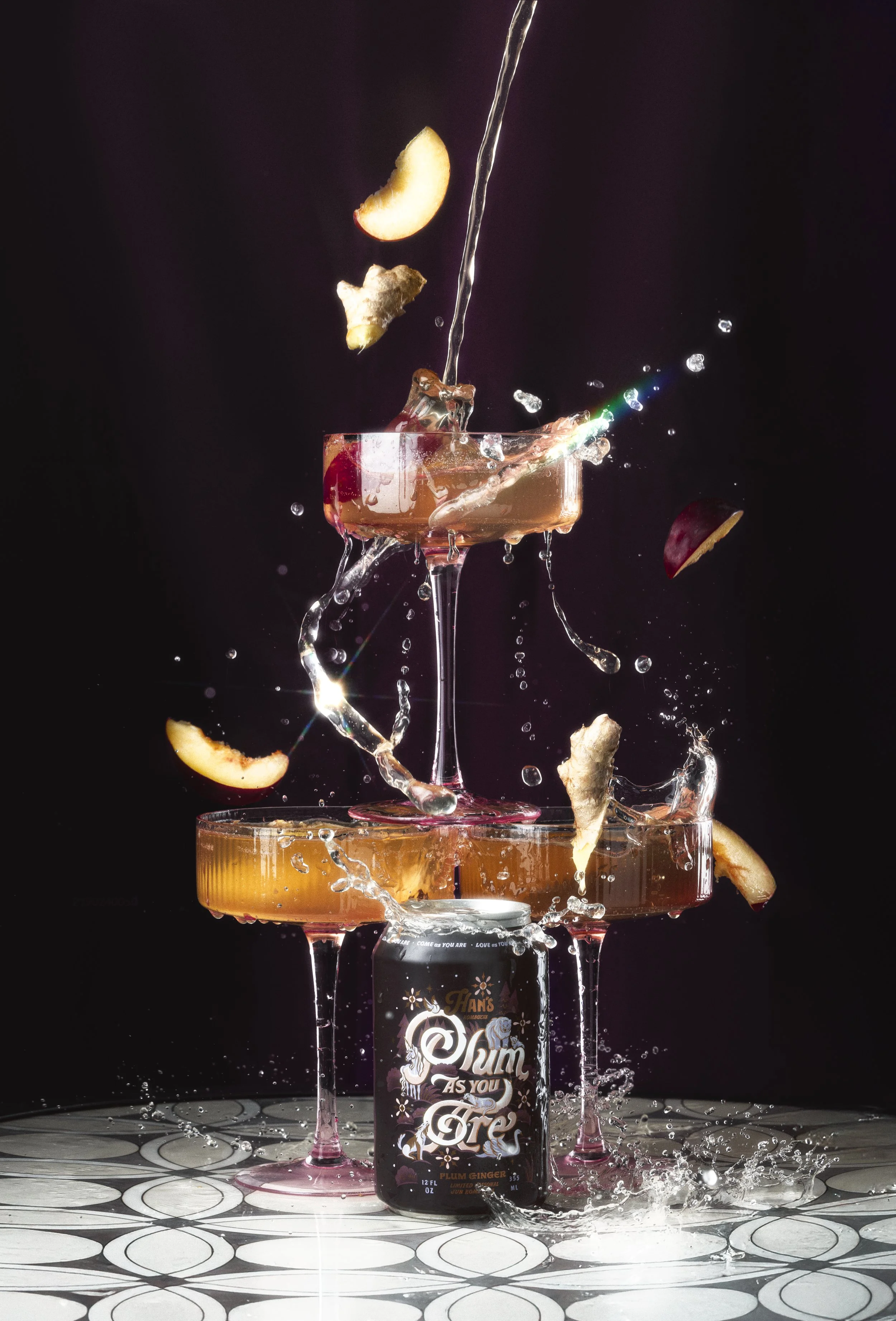 A cocktail glass with a pink drink is splashed with water, and pieces of apple and ginger are falling into it. A bottle of rum labeled 'Rum as You Feel' is in front of the glass. The scene is lit against a dark background with a patterned surface und