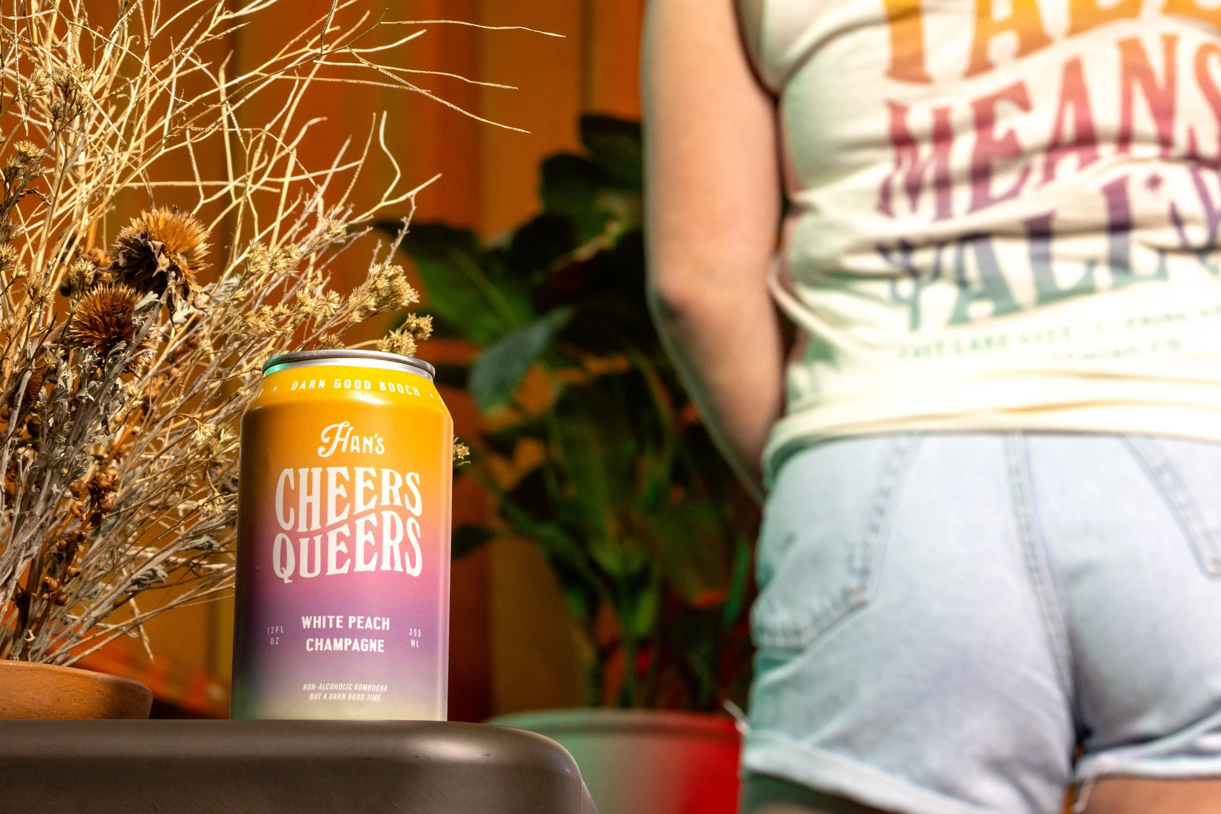 A colorful can of Cheers Queers non-alcoholic bubbly with a gradient pink, purple, and teal design placed on a table next to dried plants. In the background, a person wearing a light-colored shirt and denim shorts is partially visible.