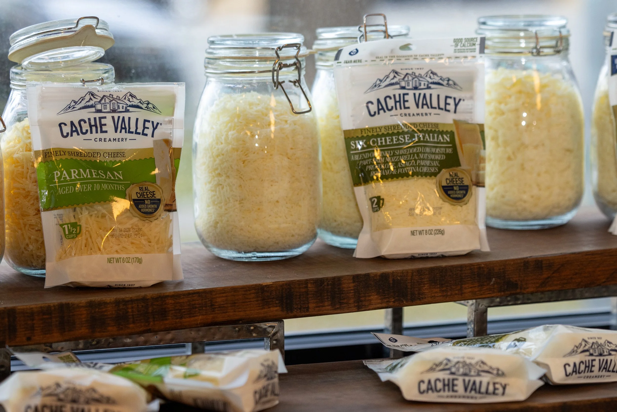 Clear glass jars filled with shredded cheese on a wooden shelf, with cheese packages labeled 'Cache Valley' and 'Finely Shredded Cheese' in front.