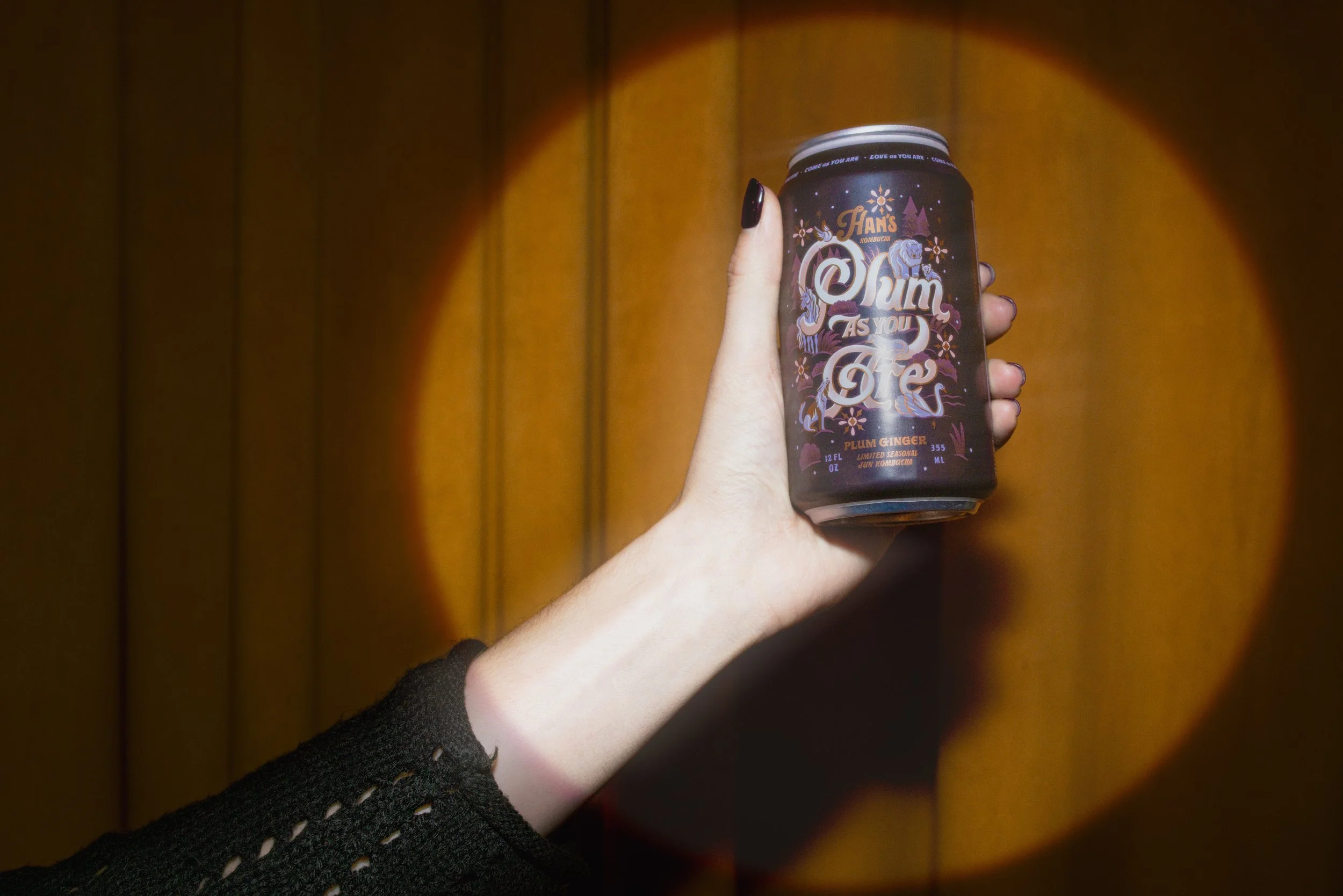 Hand holding a can of Han's Kombucha with purple and black label featuring unicorns, flowers, and the phrase 'Plumb As You Are'