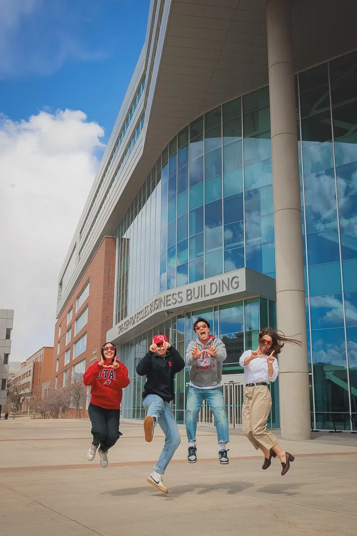 Four young people jumping in front of a modern glass building labeled 'Graduate & Continuing Business Building' on a bright, partly cloudy day.