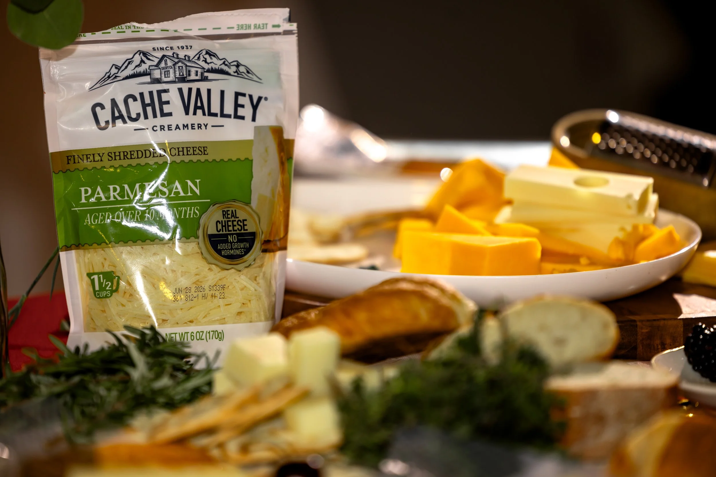 A bag of Cache Valley finely shredded Parmesan cheese in the foreground with a plate of sliced cheese, a cheese grater, and food items in the background during food preparation.