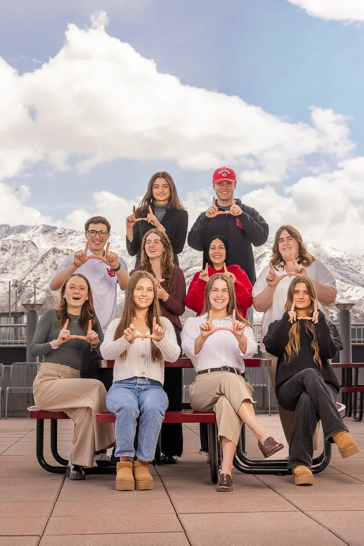 Group of young adults posing outdoors, making heart shapes with their hands, with mountains and clouds in the background.