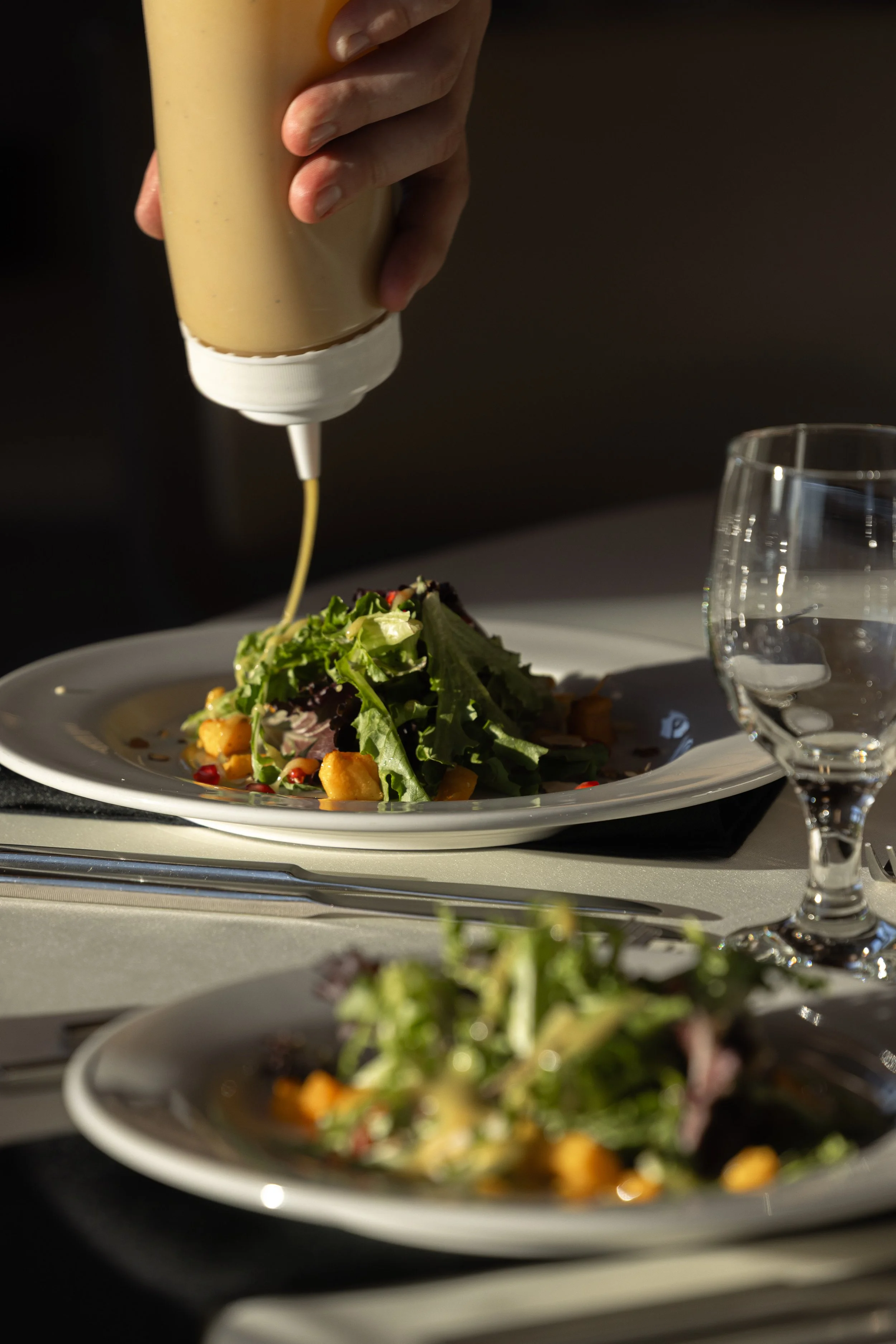 A hand holding a squeeze bottle pouring creamy dressing onto a salad with greens, vegetables, and croutons, on a white plate, with a glass of water nearby on a dining table.