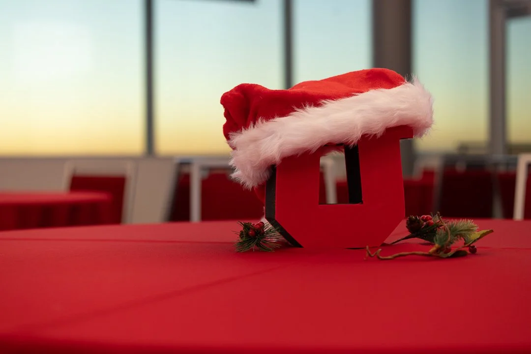 A table decorated for Christmas with a red Santa hat on top of a red gift box shaped like the letter U, surrounded by small pine branches with red berries.