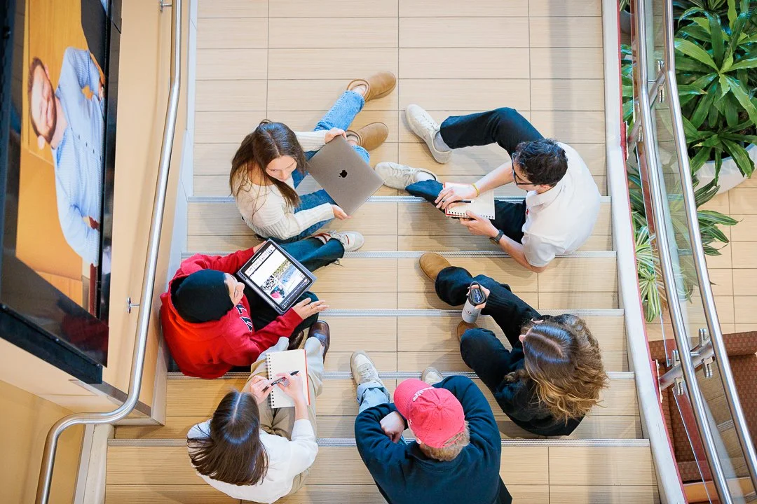 Group of six young people sitting on wooden stairs in a circle, using laptops and tablets.