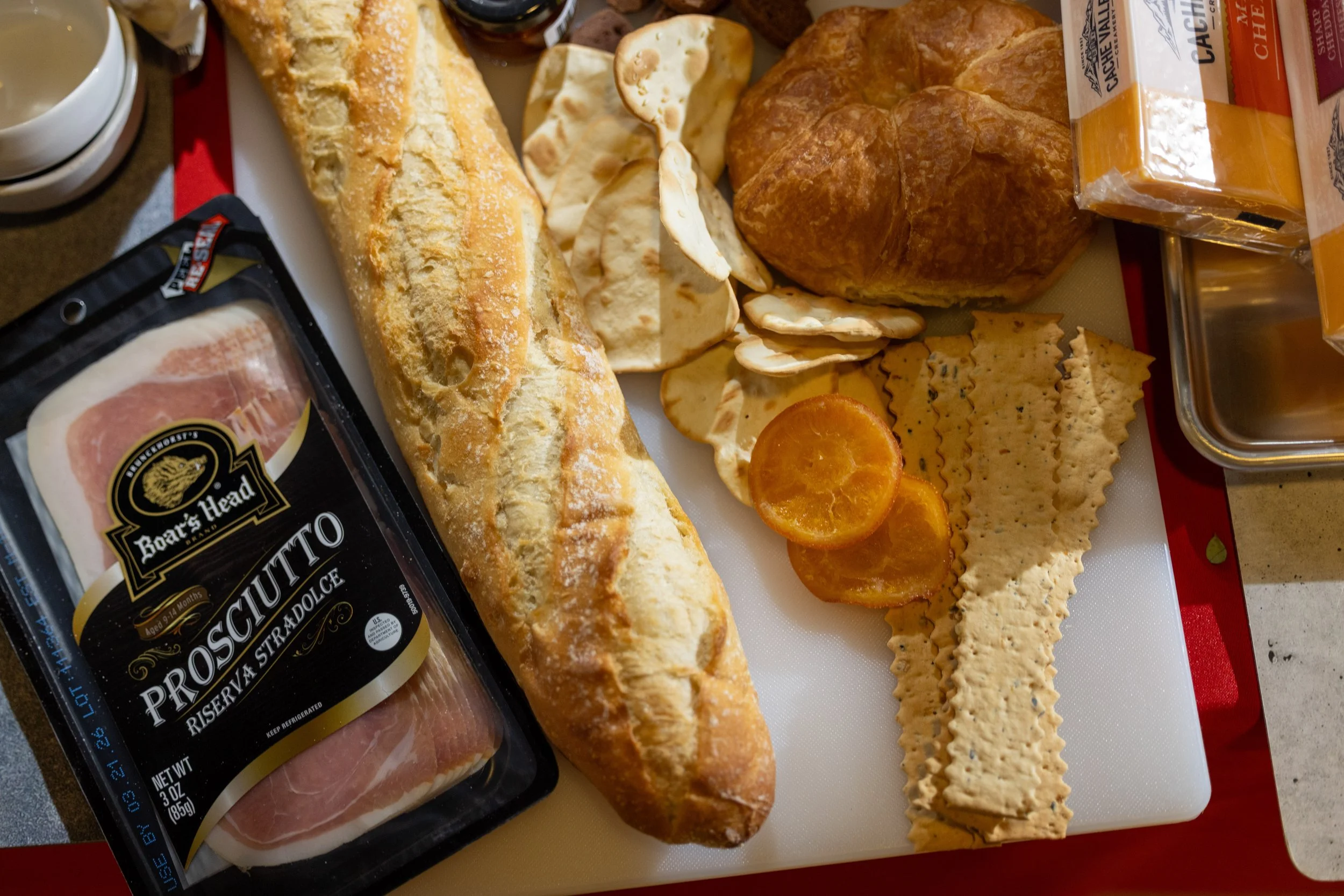 A spread of breakfast foods including a loaf of French bread, sliced cheese, sliced ham, sliced salami, a cooked egg, and prepared breakfast chips, with a jar of orange sauce and two cups on a tray.