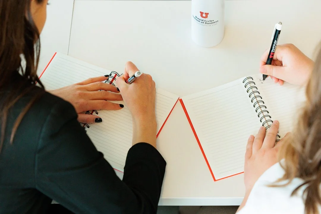 Two women sitting at a white table writing on lined spiral notebooks with black markers, with a white mug with red and black logo in the background.