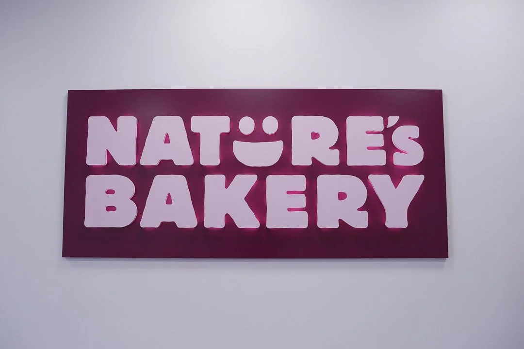 Pink sign with white text reading 'NATURE'S BAKERY' on a white wall.