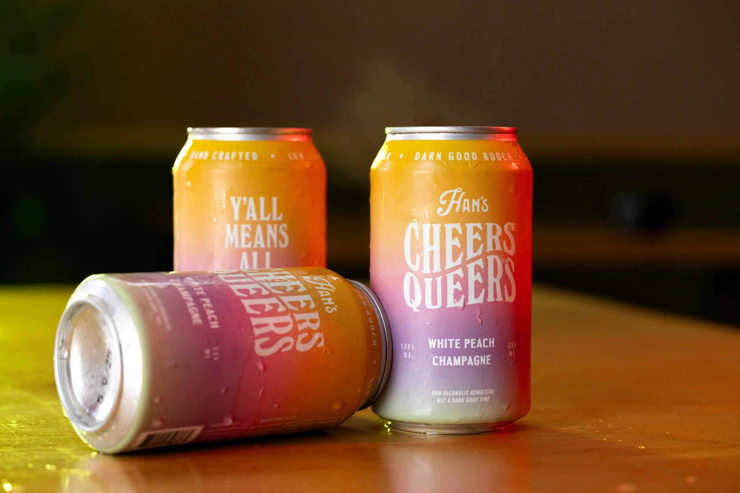 Three cans of sparkling beverages on a wooden surface with droplets of condensation. Two cans are standing upright, and one is lying on its side. The cans are pink, orange, and white with text and branding visible.