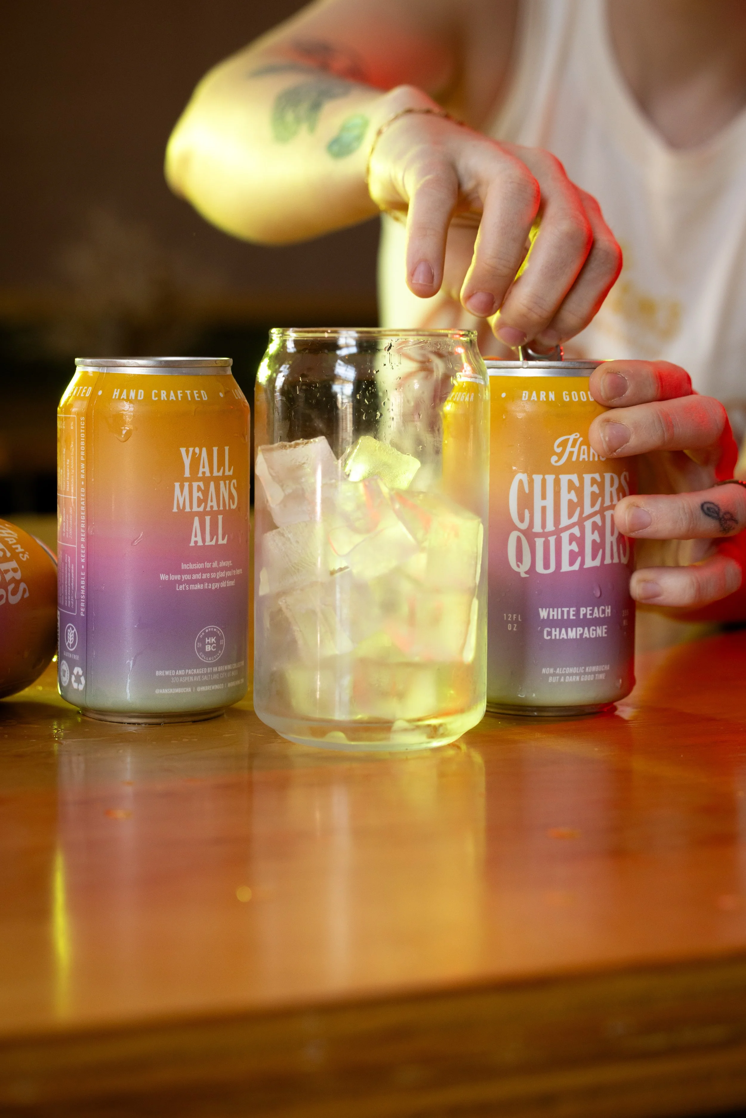 Hand holding a yellow can of Cheers Queers beer, with another can of Cheers Queers beer and a glass of a yellow-colored drink with ice on a wooden table.