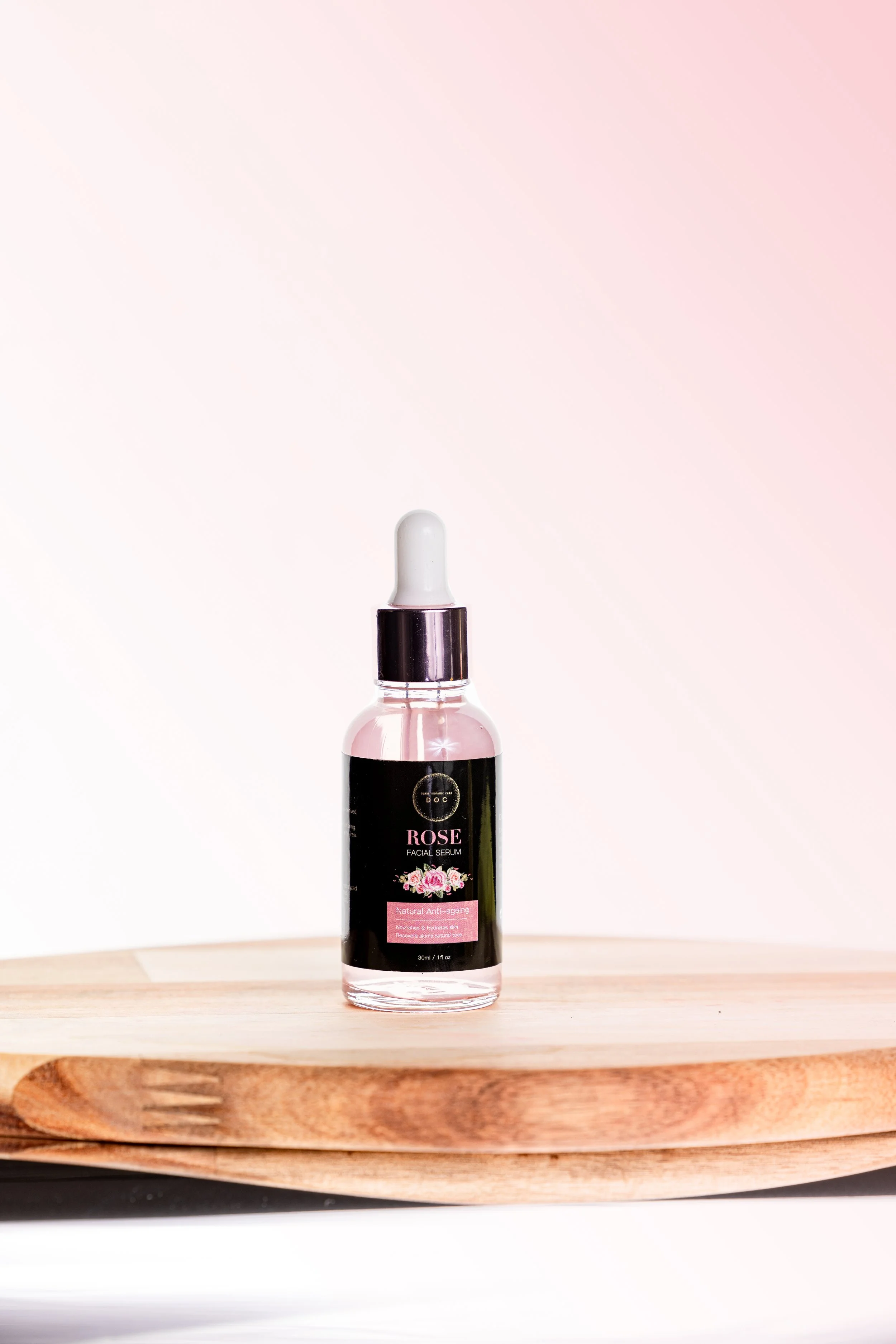 A clear glass bottle with a dropper cap containing rose facial serum, placed on a wooden surface with a soft pink gradient background.