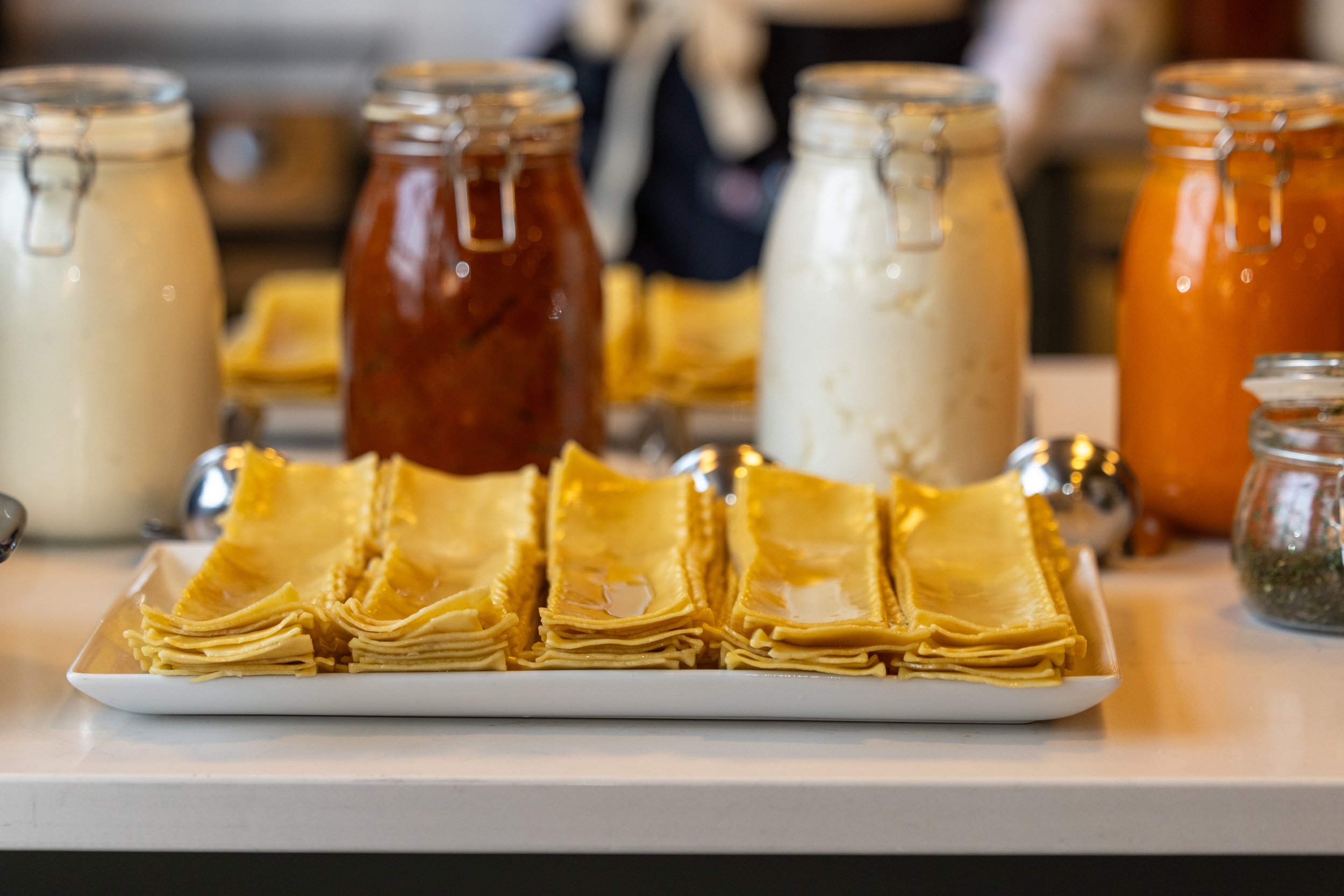 A white rectangular platter holds neatly arranged uncooked lasagna sheets. In the background, there are jars of white sauce, red sauce, and orange sauce, along with a small jar of herbs, all arranged on a table. The setting appears to be a kitchen or