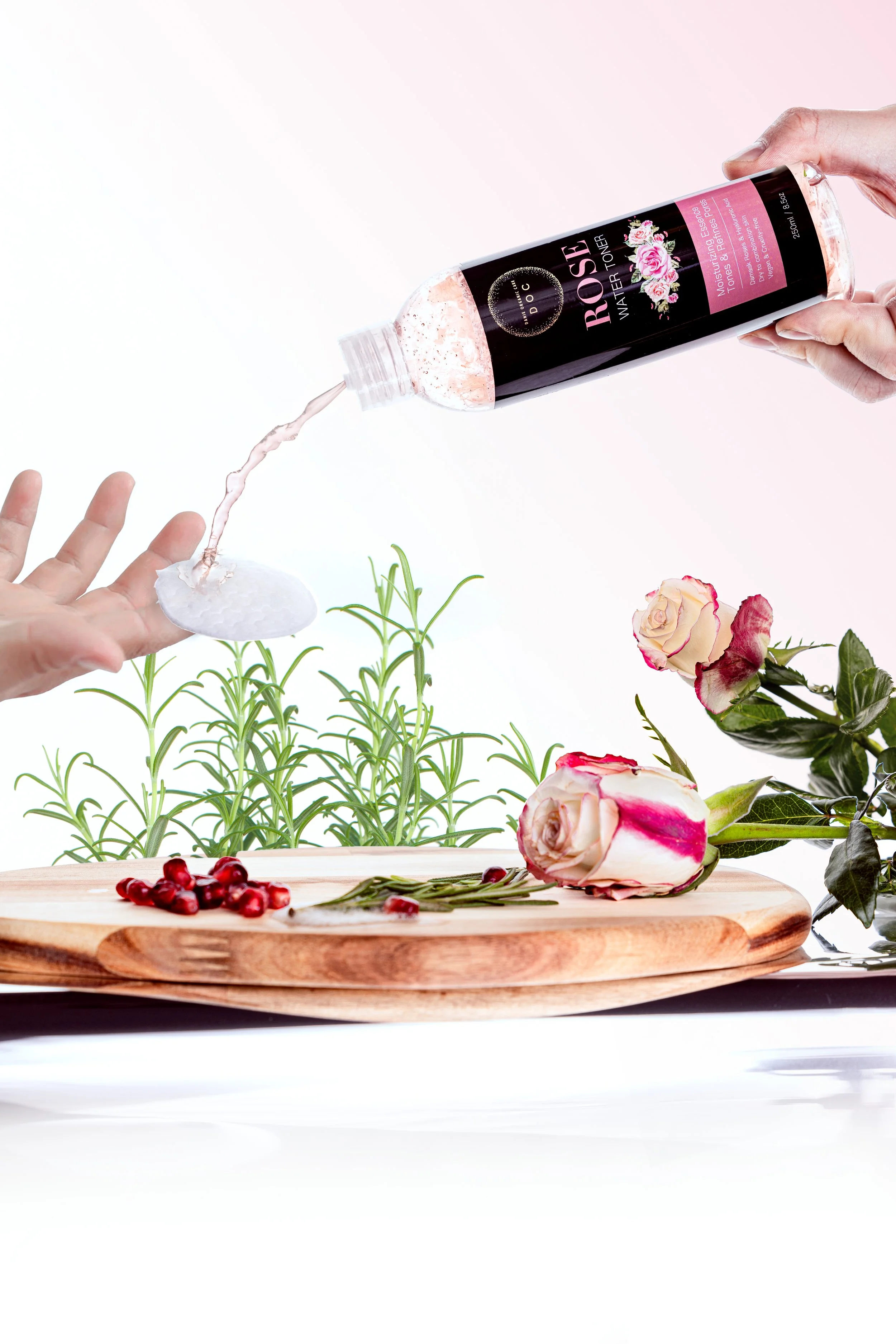 A person pouring rose water from a bottle onto their hand with roses, pomegranate seeds, and herbs on a wooden tray, and various flowers and greenery in the background.