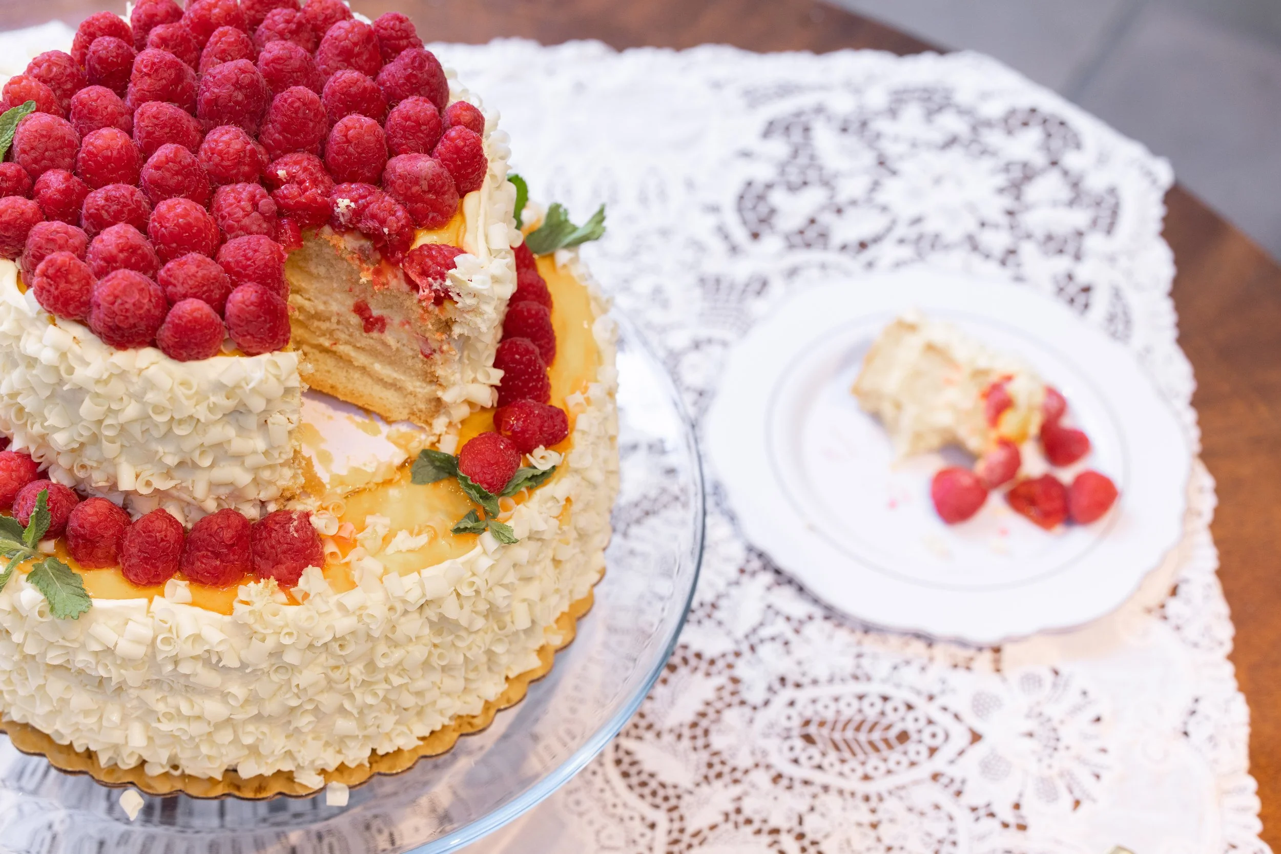 A round, decorated cake with vanilla frosting, topped with raspberries, mint leaves, and white chocolate curls, placed on a glass cake stand on a lace tablecloth with a plate of a partially eaten slice.