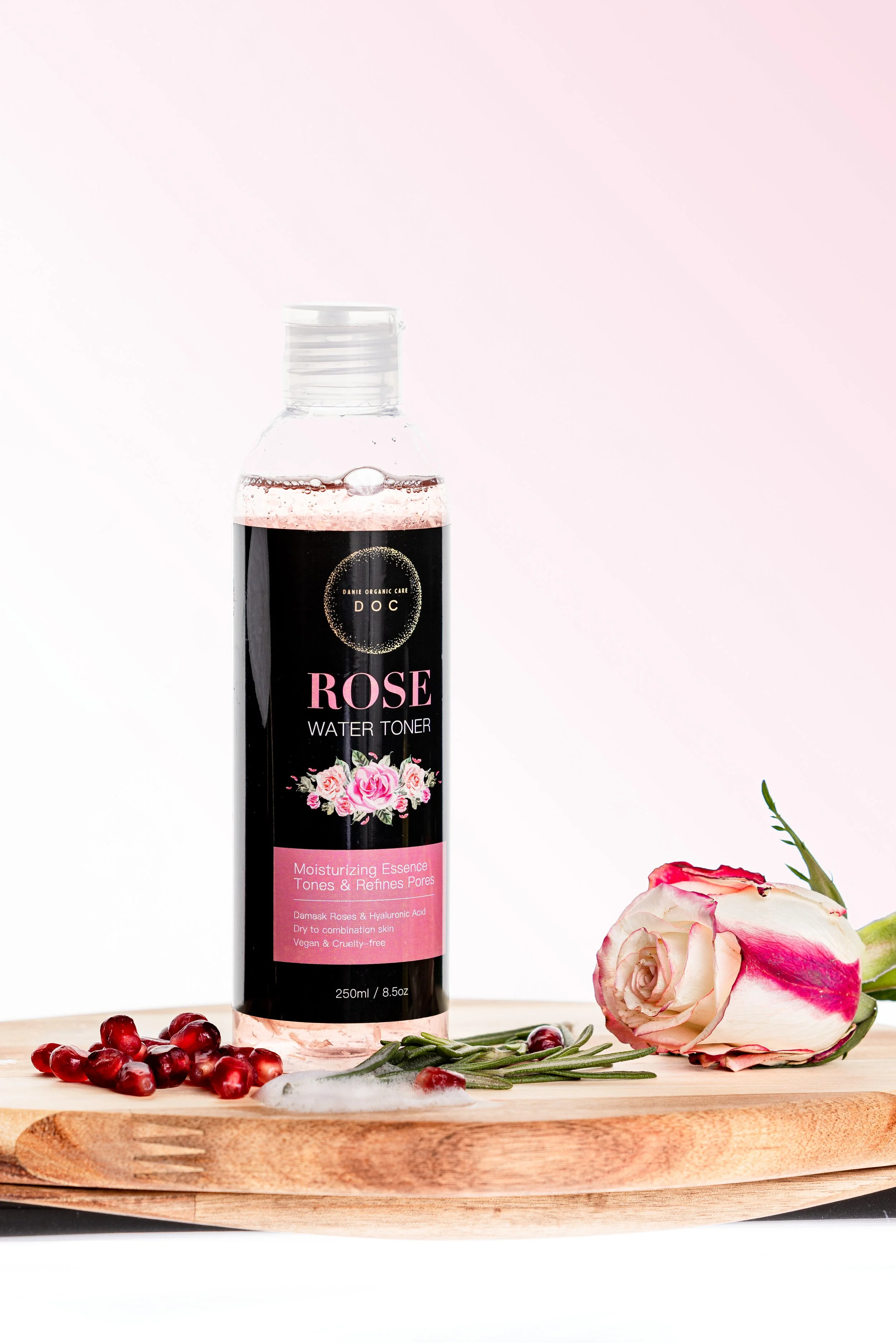 A bottle of rose water toner on a wooden surface surrounded by pomegranate seeds, a pink and white rose, and green herbs, with a light pink and white background.