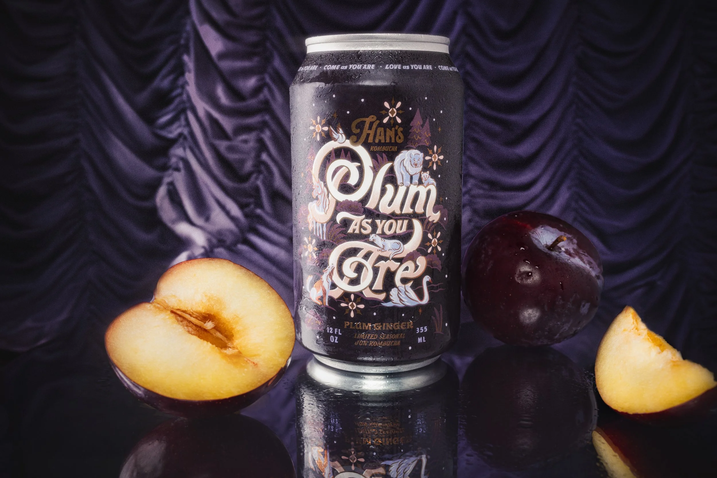 A can of Han's kombucha with a dark purple and black design featuring a lion and mystical patterns, placed on a reflective surface surrounded by halved peaches and a whole dark red apple, with a purple velvet background.