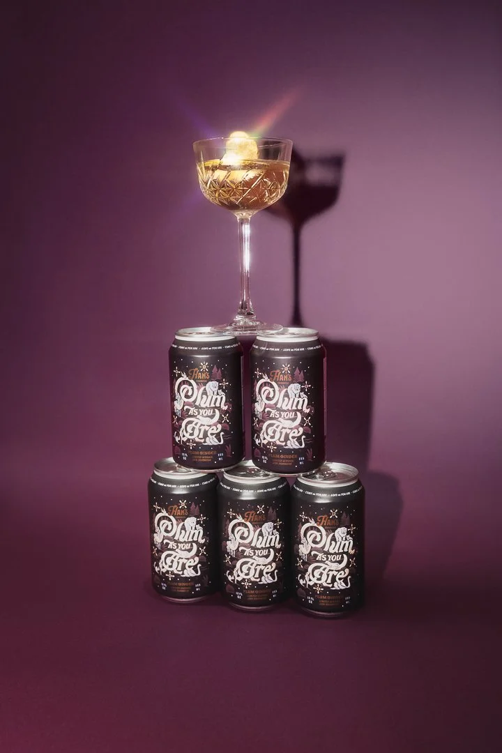 A pyramid of five cans labeled 'Plumb As You Are' on a purple background, topped with a cocktail glass containing a drink and a Plumb slice, casting a shadow on a Plumb colored wall.
