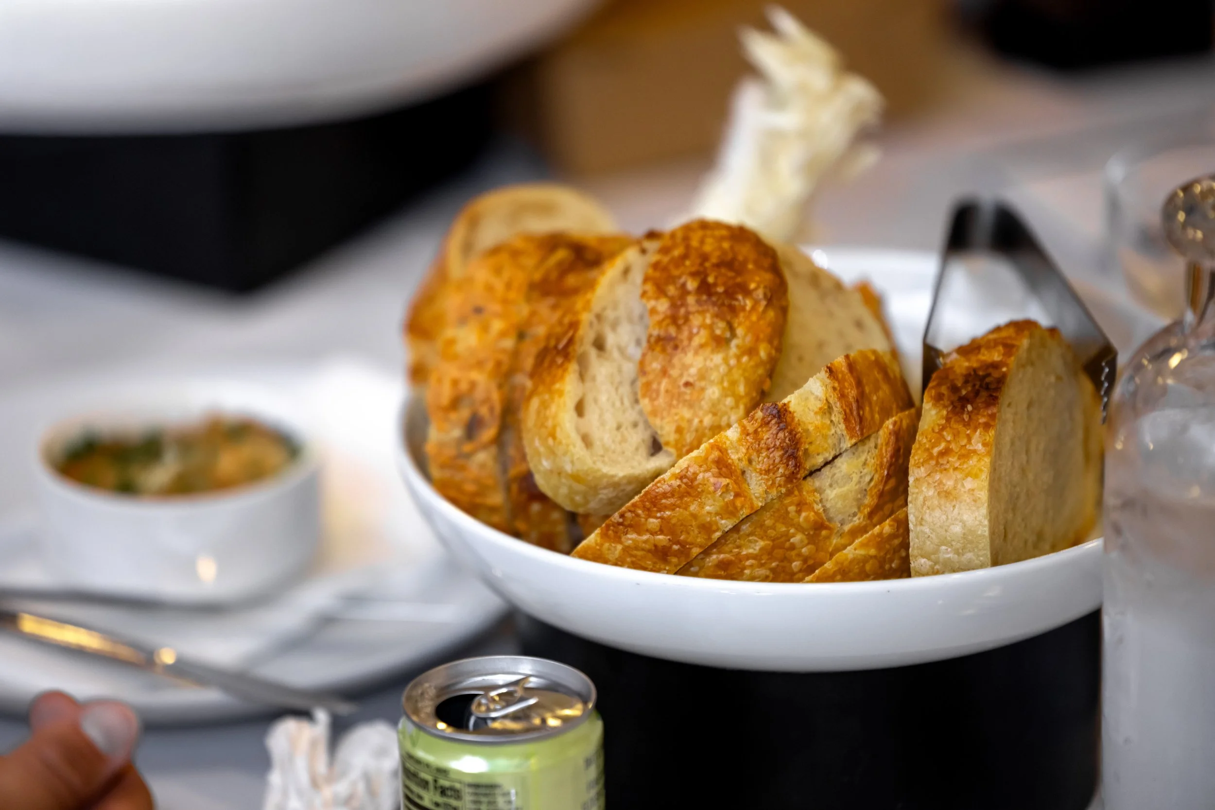 Close-up of sliced garlic bread in a white bowl with a person holding a soda can in the foreground, and other dishes slightly blurred in the background.