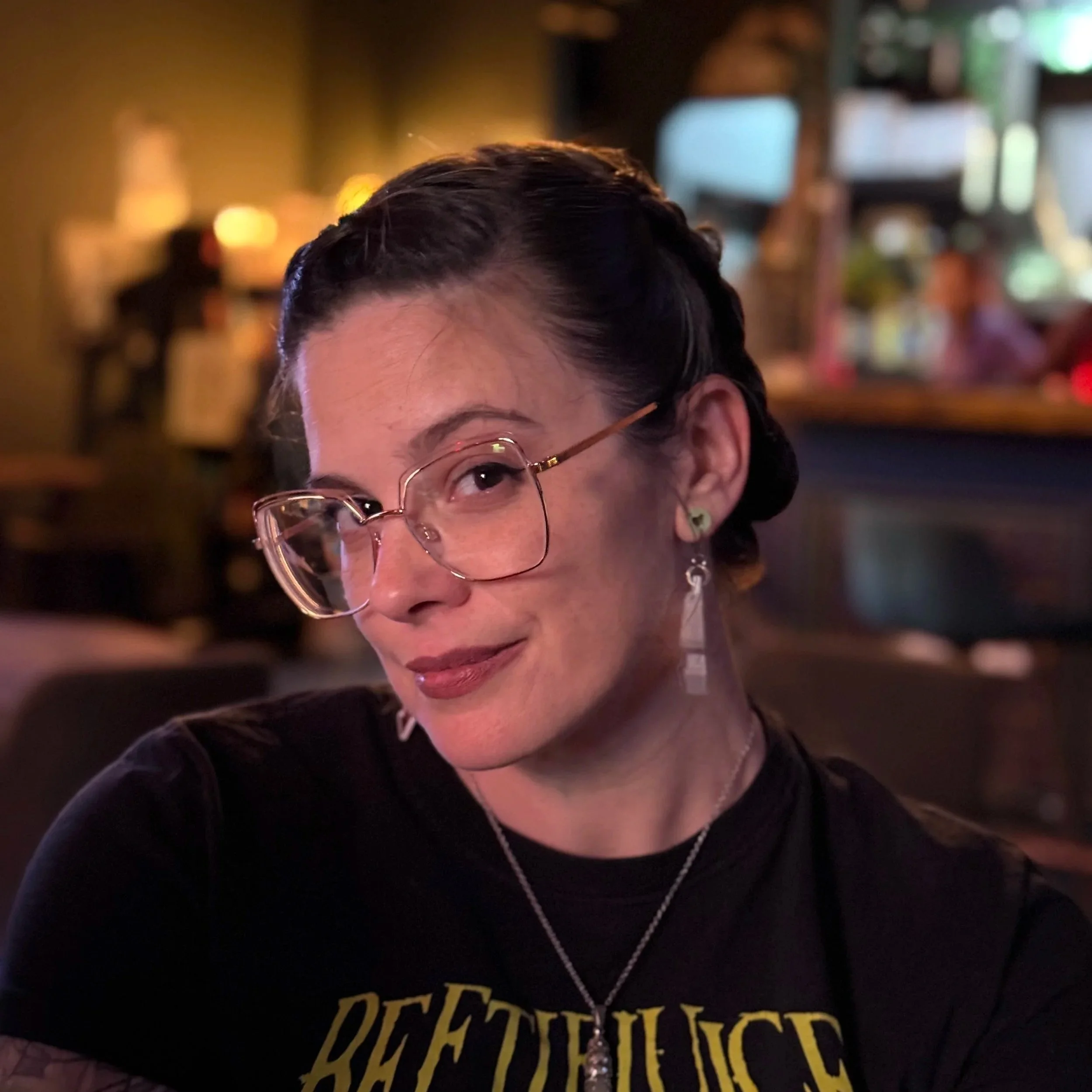 Close-up of a woman with short dark hair, glasses, and earrings, wearing a black shirt with yellow text, in a dimly lit indoor setting with warm lighting.