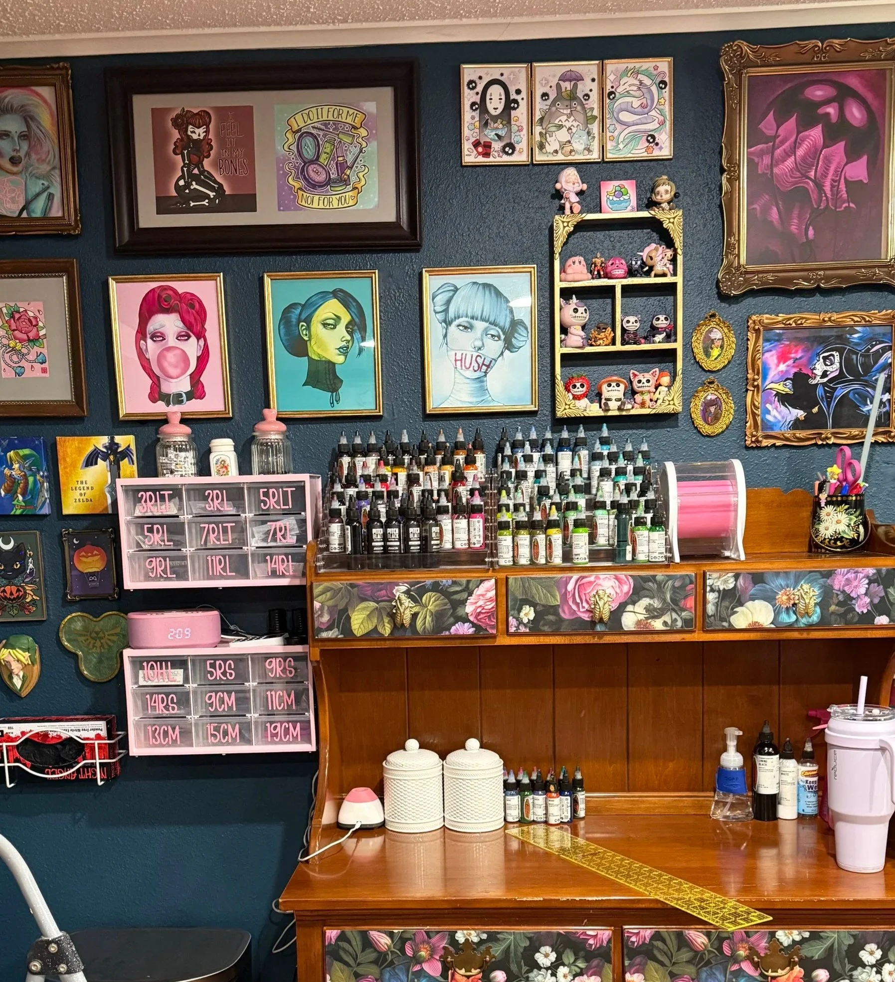 Wall decorated with various art prints, including characters from popular media and stylized illustrations. A shelf with small figurines and bottles of tattoo ink, along with other tattoo supplies a wooden hutch.