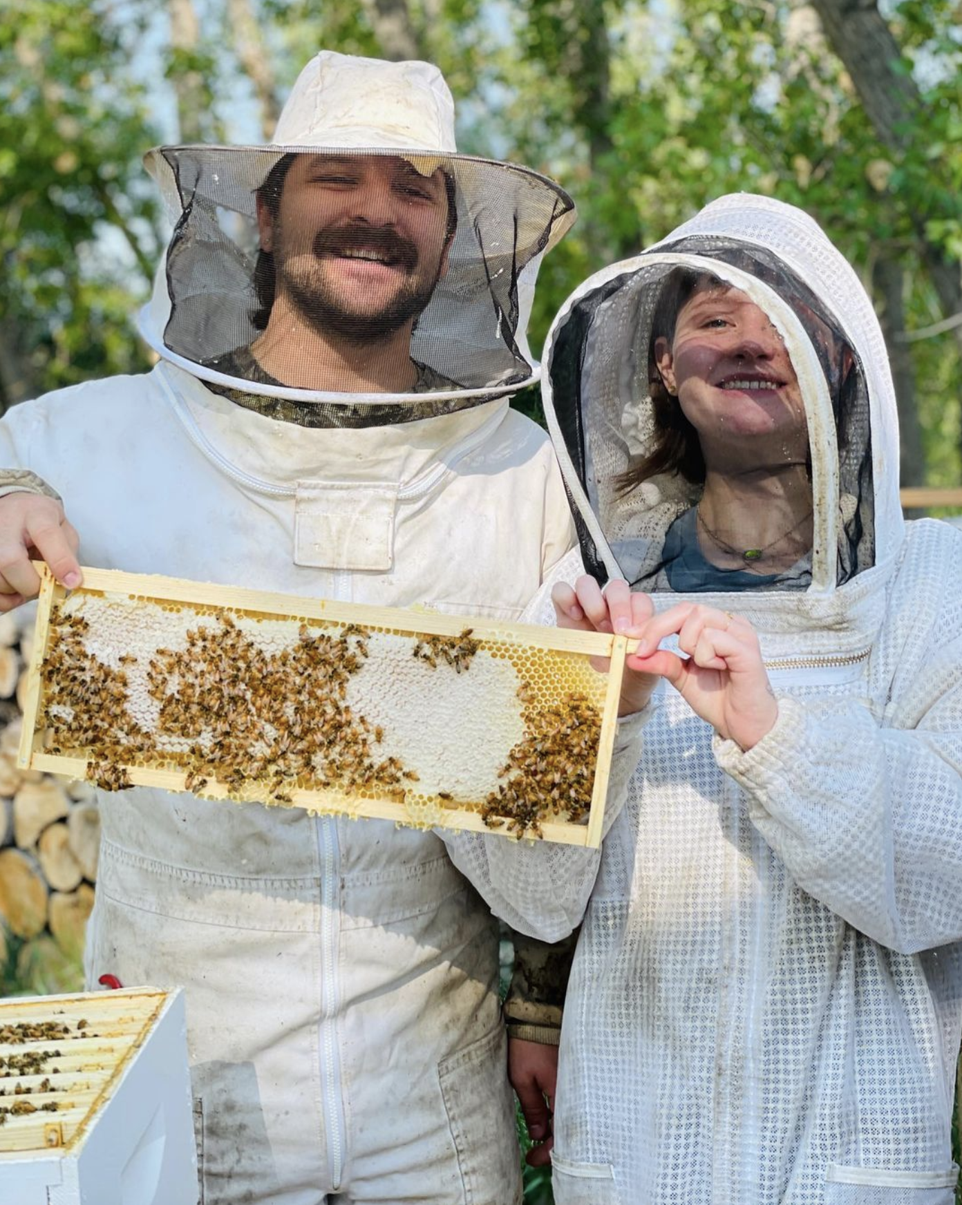 Two people in beekeeping suits holding a frame of bees and honey comb.