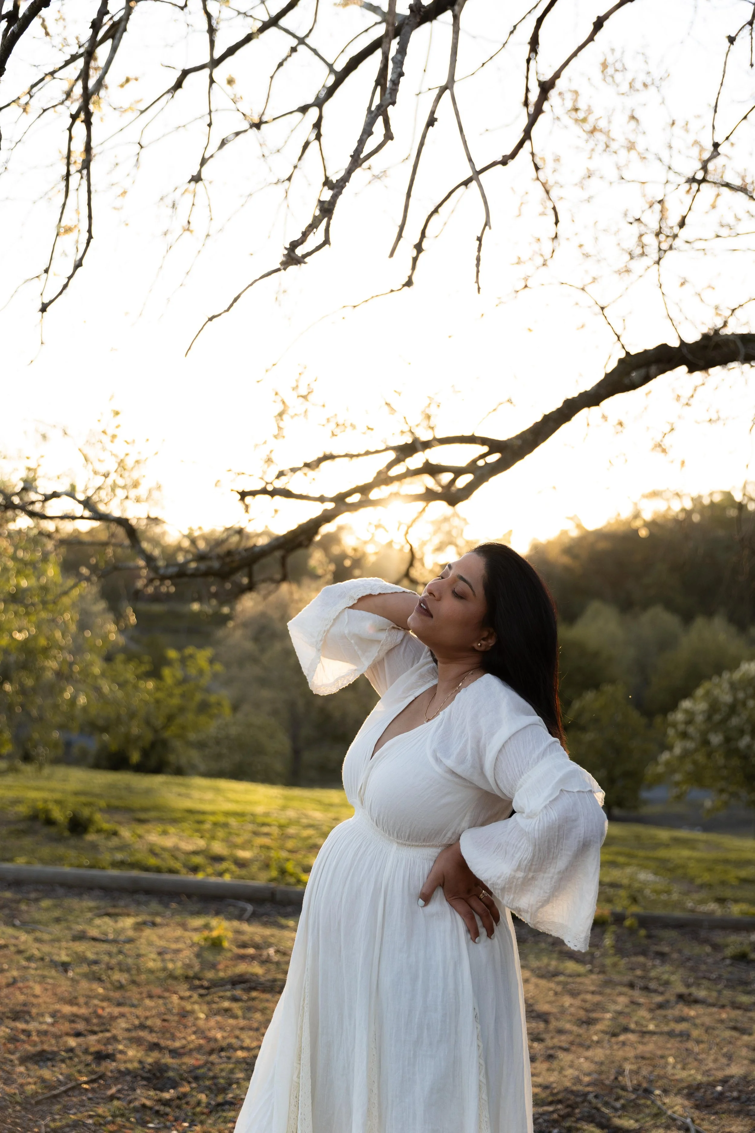 A pregnant woman in a white dress standing outdoors at sunset, with her hand on her hip and eyes closed, smiling softly. Beautifully sunlit from behind. goddess vibes, earthy mumma