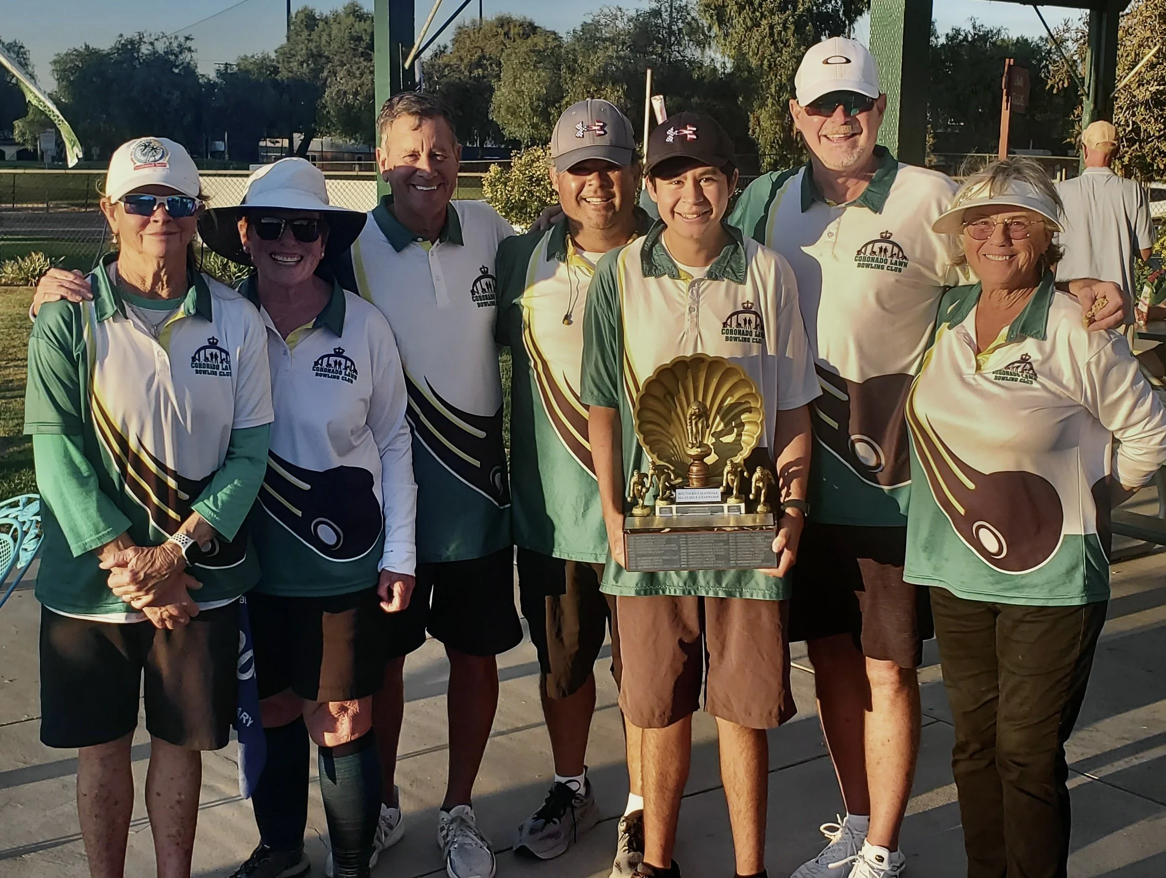 Group of eight people in matching bowling shirts standing outdoors, holding a trophy, celebrating after a bowling event.