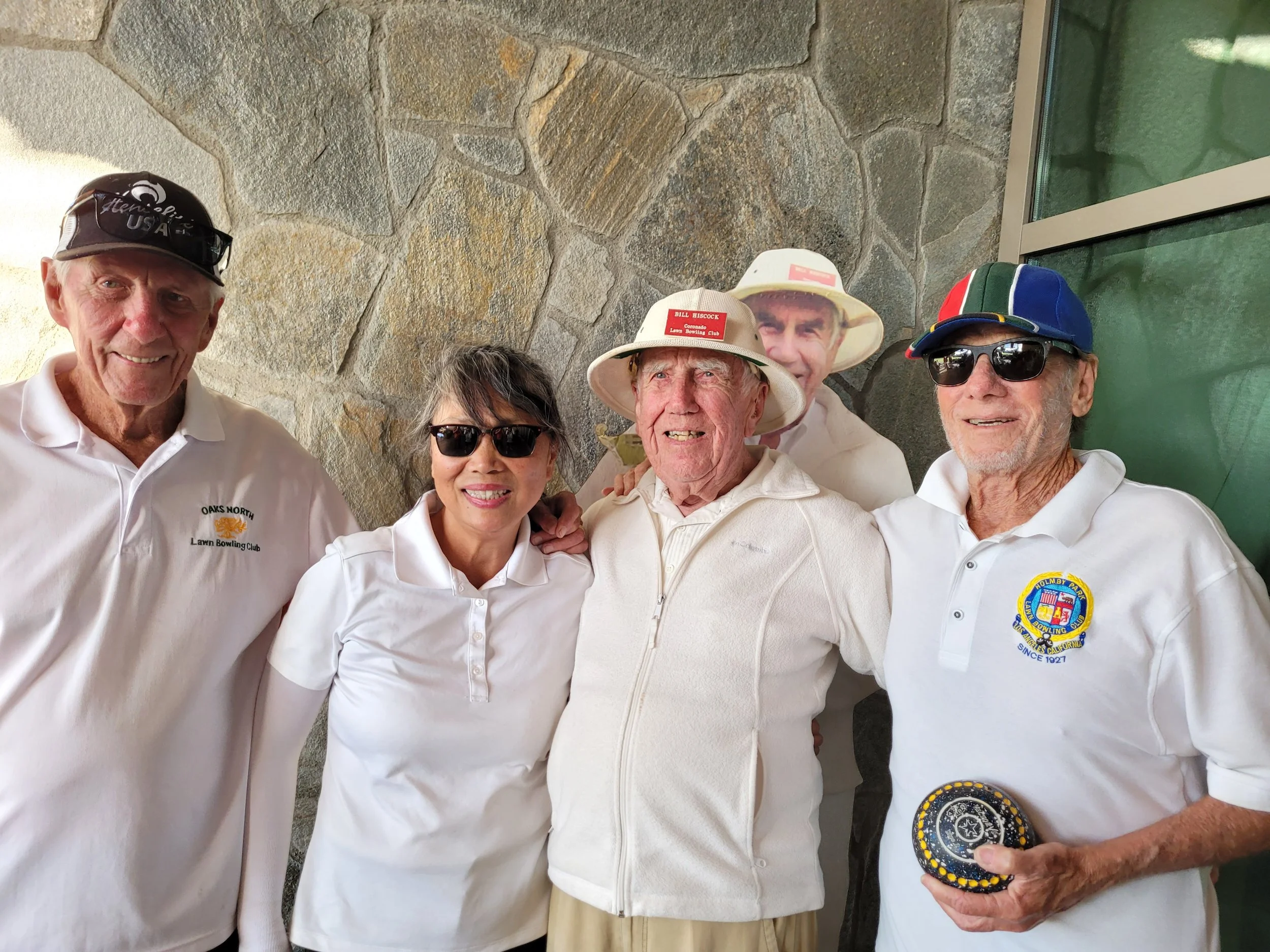 Group of five elderly adults, four men and one woman, standing together in casual sportswear, some wearing sunglasses and hats, smiling at the camera, with a stone wall in the background.