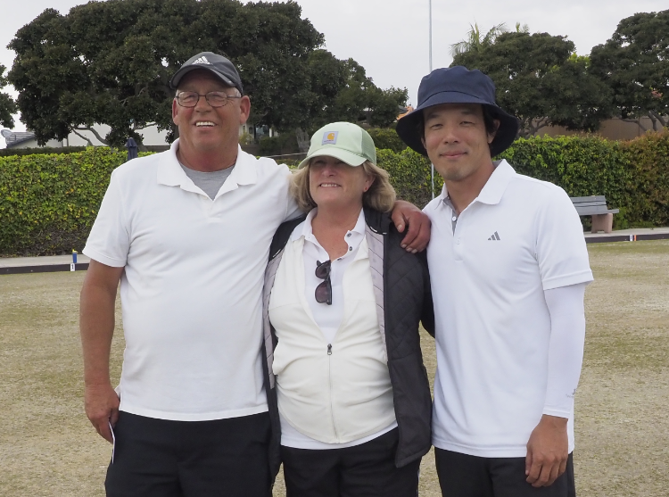 Three people standing outdoors on a grassy area, smiling, with trees and a bench in the background. Two are men, one woman, all wearing casual hats and white shirts.