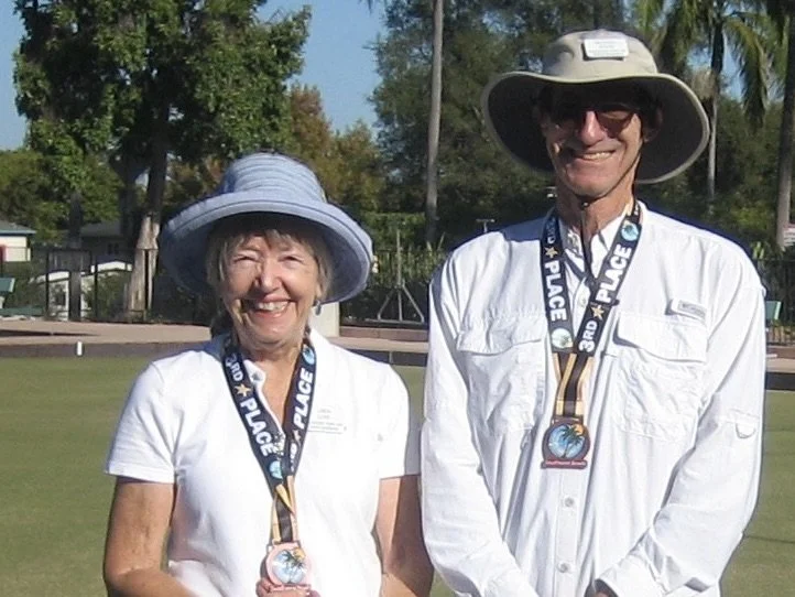 A smiling elderly woman and man wearing wide-brimmed hats and medals, standing outdoors on a grassy field with trees in the background.