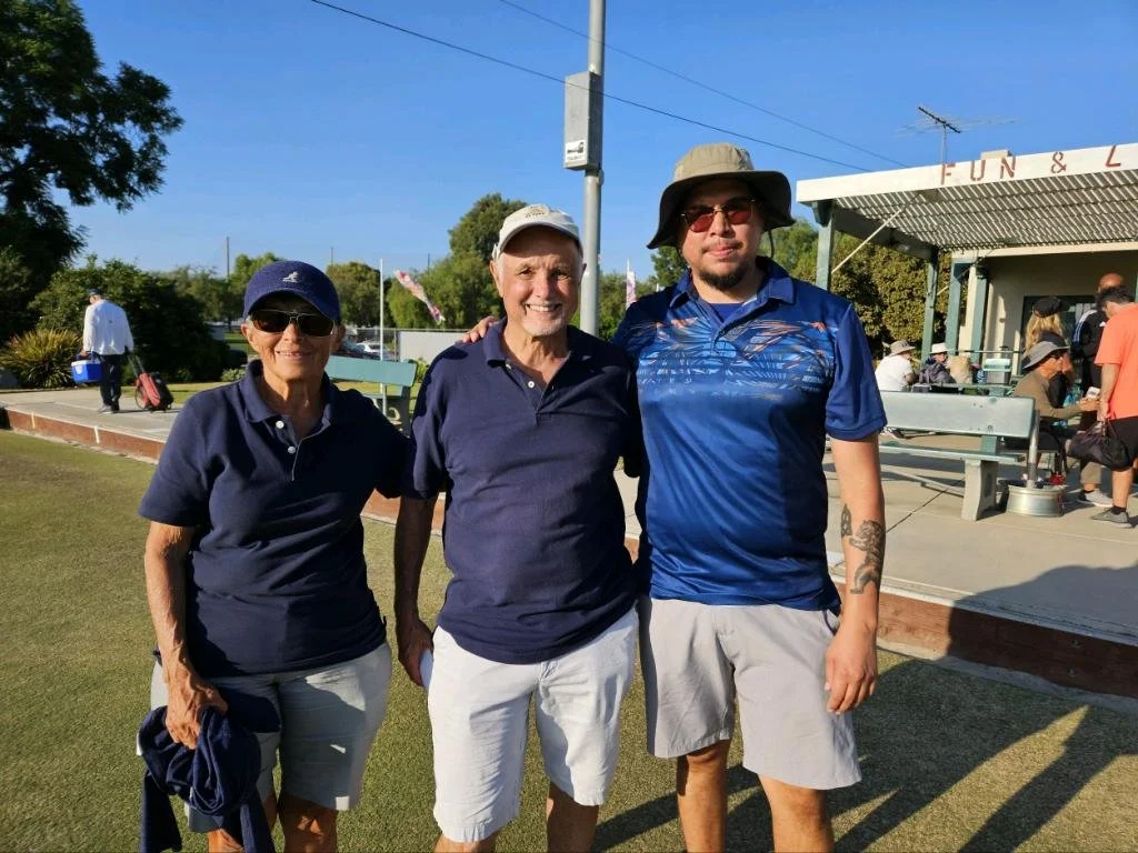 Three people smiling and standing outdoors near a golf course, wearing casual summer clothing including shorts, hats, and sunglasses.