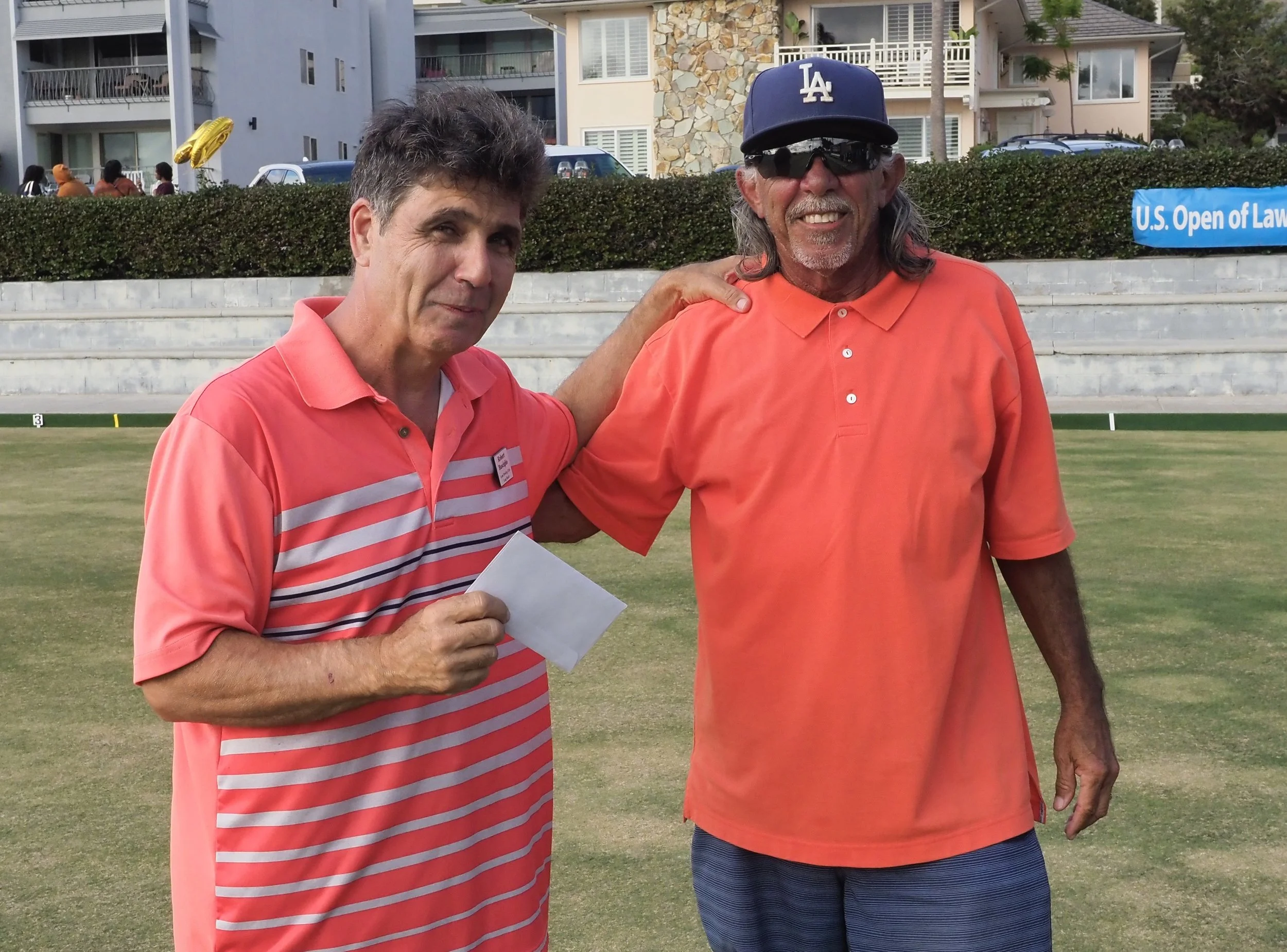 Two men on a golf course removing their hats and smiling. One man has his arm around the other. There are residential buildings and a blue sign that reads 'U.S. Open of Law' in the background.