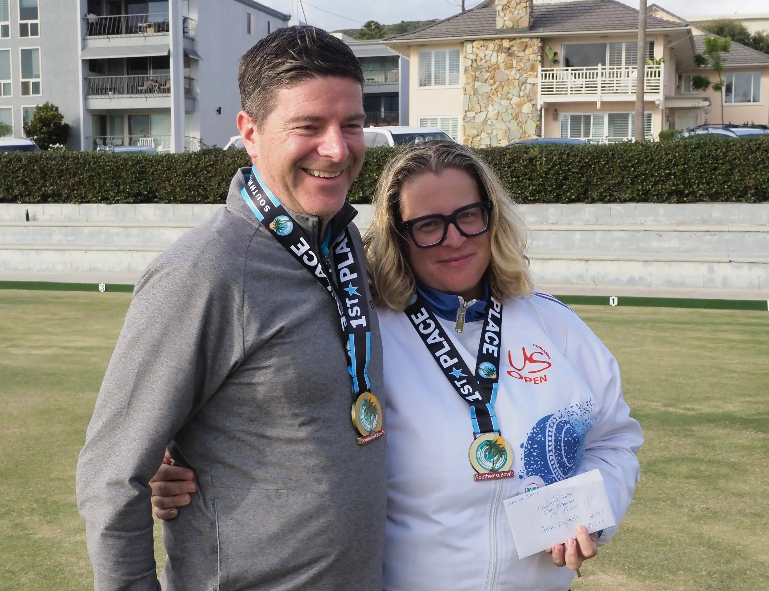 Two people smiling with medals around their necks on a lawn, holding a check, with houses and shrubbery in the background.