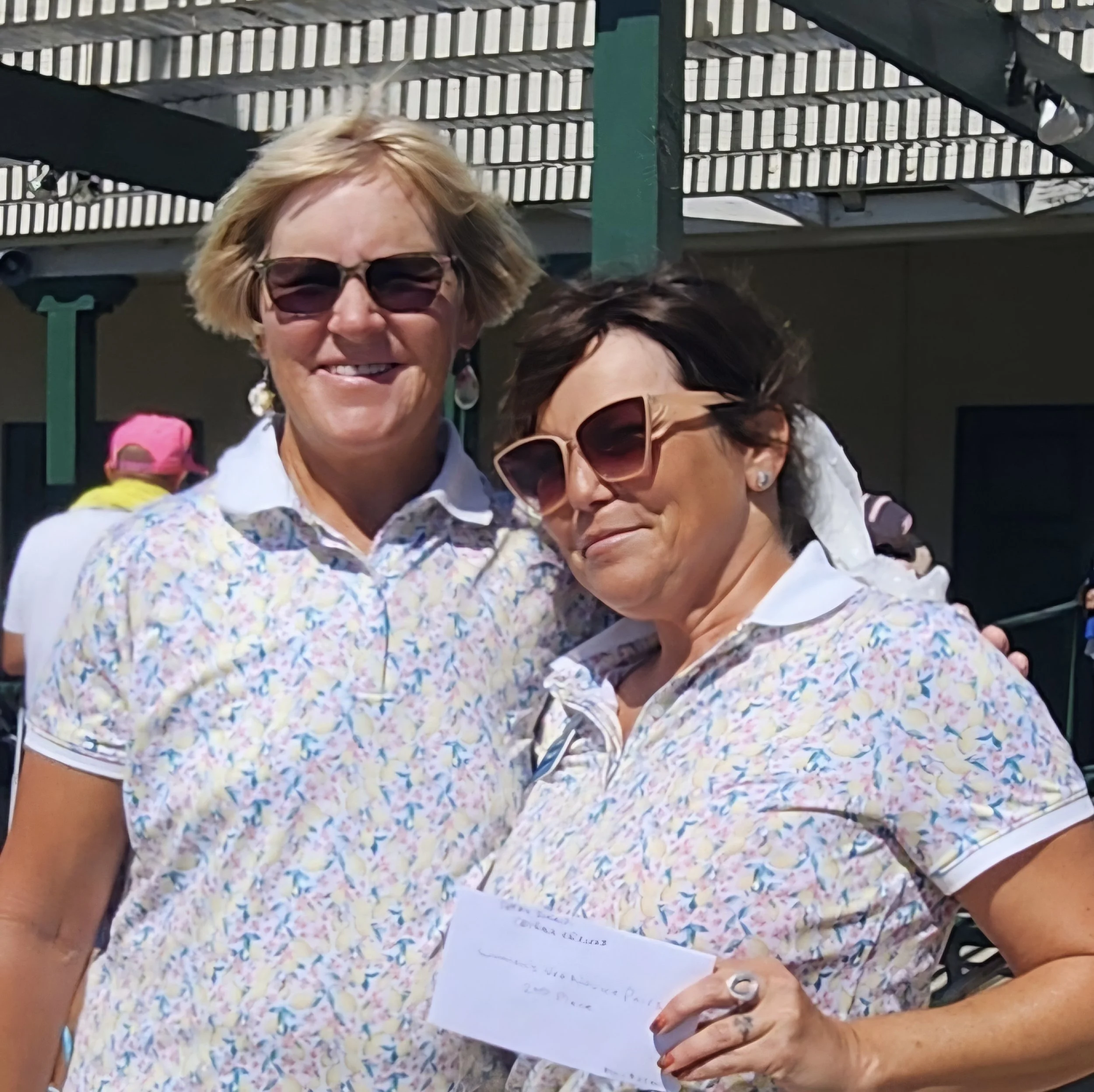 Two women wearing matching floral shirts and sunglasses, posing outdoors with a sunny background, smiling at the camera.
