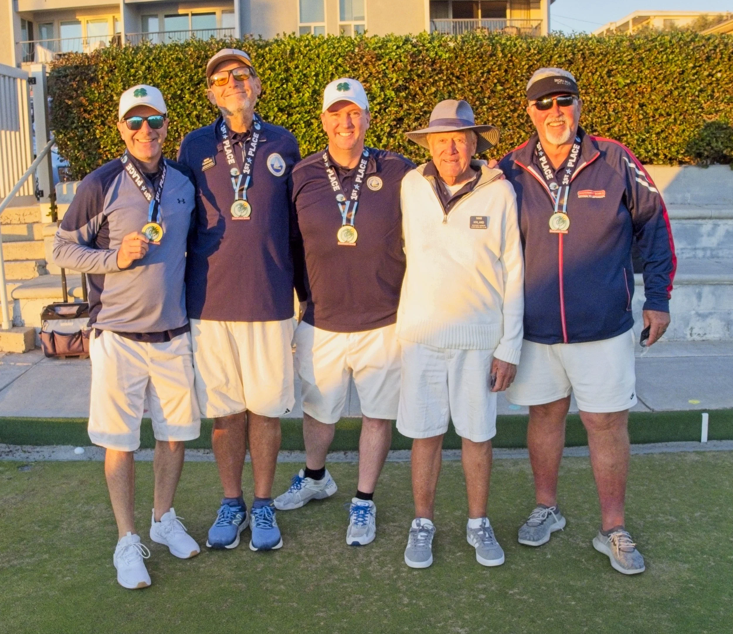 Five men standing together on a golf course, wearing medals around their necks, with a hedge and a building in the background, celebrating a golf tournament victory.
