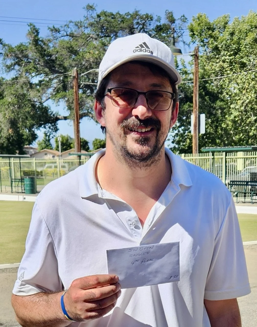 A man smiling outdoors, wearing a white Adidas cap, glasses, and a white polo shirt, holding an envelope. There are trees, a fence, and a blue sky in the background.