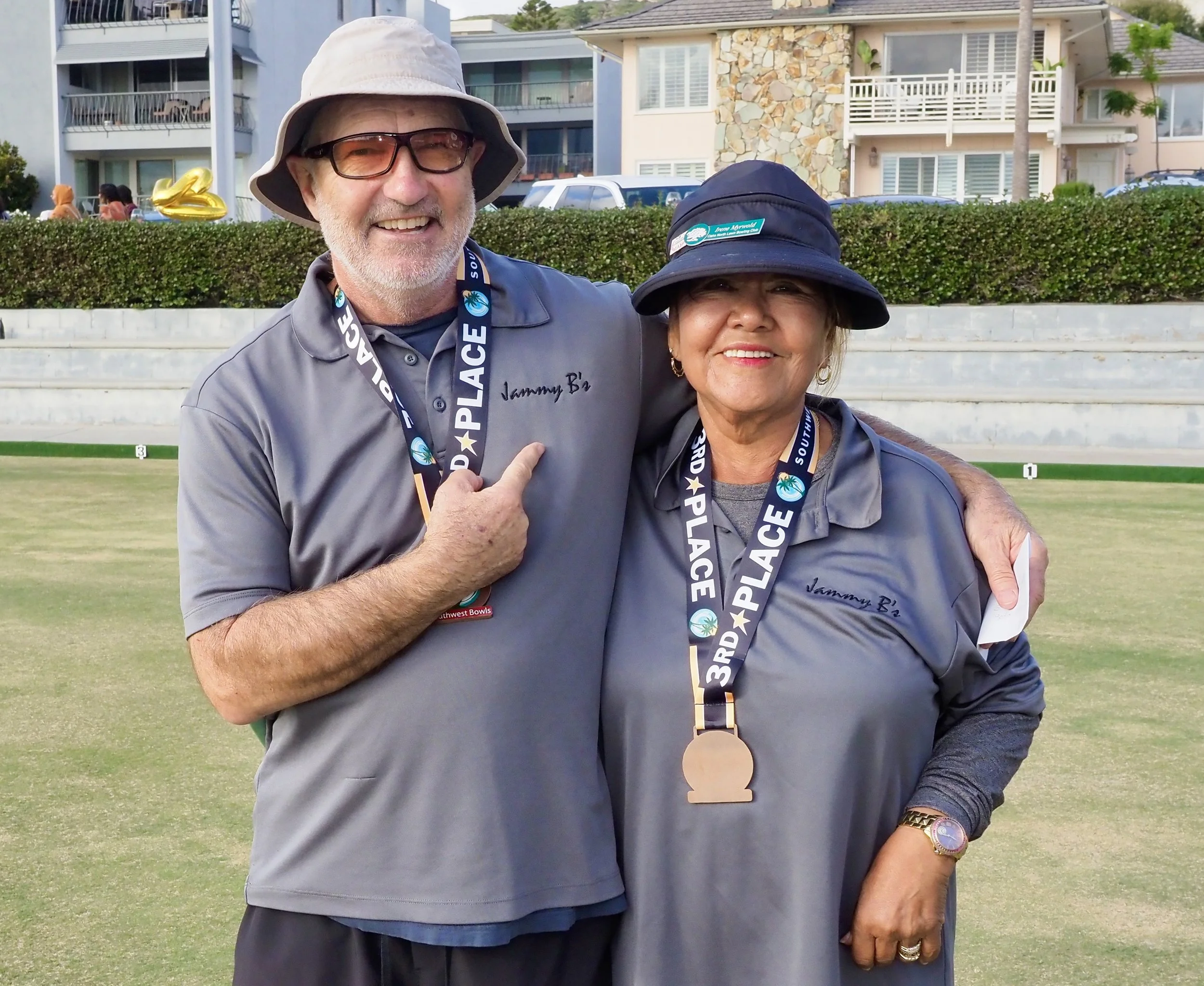 A man and a woman wearing matching gray shirts and hats, standing together on a grass field, each with a medal around their neck, smiling at the camera. The man is pointing to his shirt, and they are embracing. In the background, there are residential buildings and a small group of people.