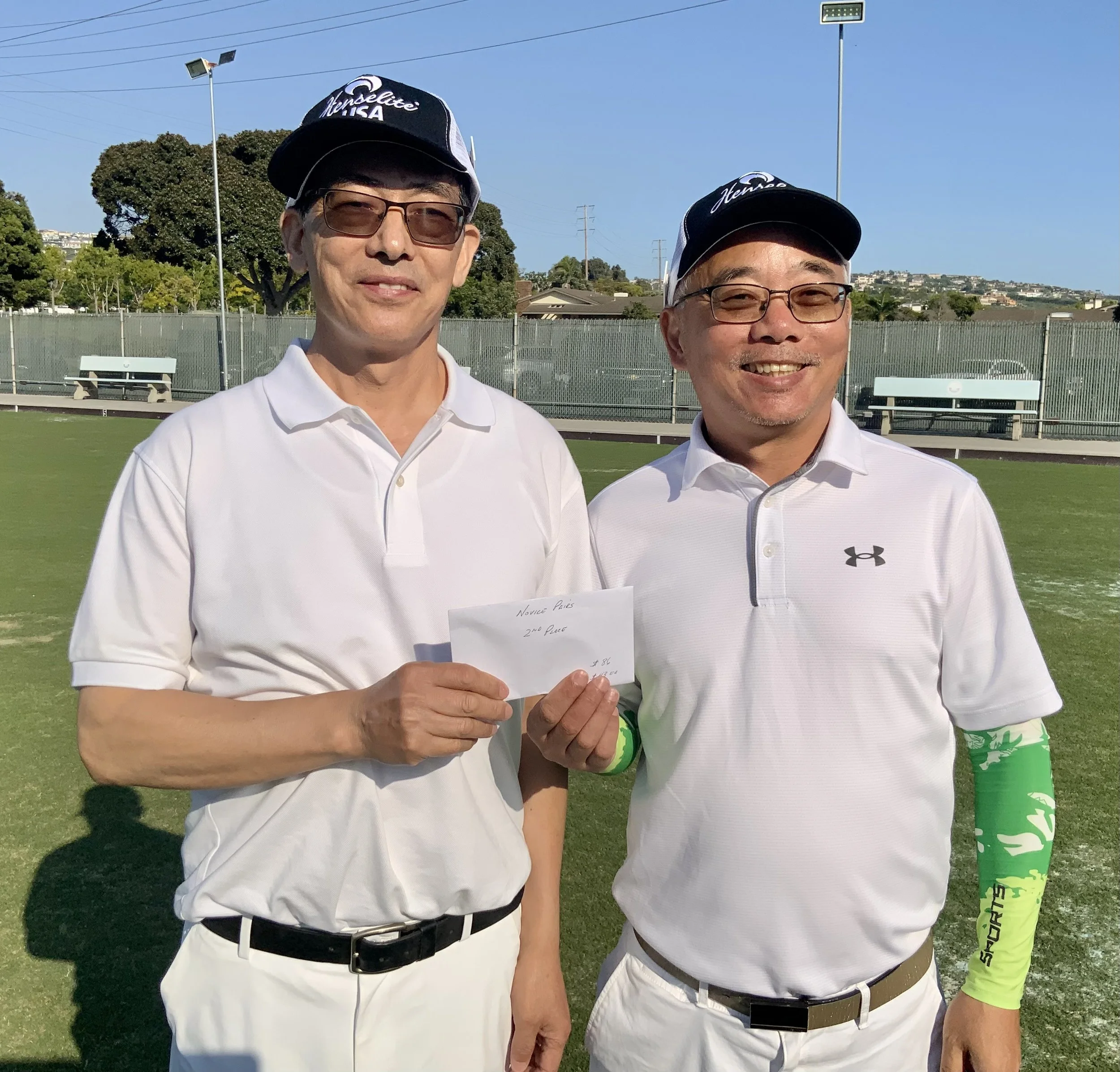 Two men in white golf attire standing on a golf course, one holding an envelope that says 'Nomura Paris, 2nd Place,' both smiling and wearing caps and sunglasses.